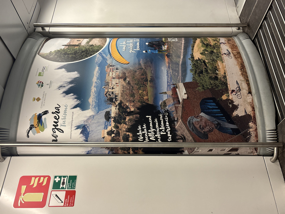 The image depicts the interior of a public transport vehicle, likely a train car, located in Sant Quirze del Vallès, Spain. A large, vertically oriented advertisement poster for "Noguera Turisme" dominates the right side of the frame, spanning from near the ceiling to the floor. The poster is a collage of scenic landscapes and activities, featuring snow-capped mountains, rural villages, a lake or reservoir, and lush green hills.
Within the poster's imagery:
* The top section includes a logo with a stylized bird or creature and the text "Noguera Turisme" with the website "www.turismenoguera.cat". Several smaller institutional logos, including "CONSELL COMARCAL DE LA NOGUERA" and "FGC", are present at the top right.
* A circular inset image on the upper right displays a stone building, possibly a castle or historic structure, surrounded by cypress trees.
* Activities depicted include a yellow paraglider with two figures below it, and three individuals on bicycles riding along a dirt road in a lower section.
* Key promotional terms in black text on a textured background include "Natura," "Aventura," "Allotjament," "Gastronomia," "Patrimoni," and "Experiencies" (Nature, Adventure, Accommodation, Gastronomy, Heritage, Experiences).
* A prominent illustration of an older man with a smiling expression, wearing a flat cap and a striped shirt, is situated in the lower-left quadrant of the poster.
To the left of the poster, a white interior wall displays three safety pictograms: a red square with a yellow fire extinguisher symbol (Escala de socors), a green square with a white downward staircase symbol, and a green square with a white first aid symbol (Camilla de socorro). A silver-colored metal vertical pole, likely a handrail, runs along the left edge of the poster, extending from top to bottom. The floor shows a dark, ribbed grating design on the right, transitioning to a light gray, textured surface on the left. No individuals or direct interactions are visible outside the poster.