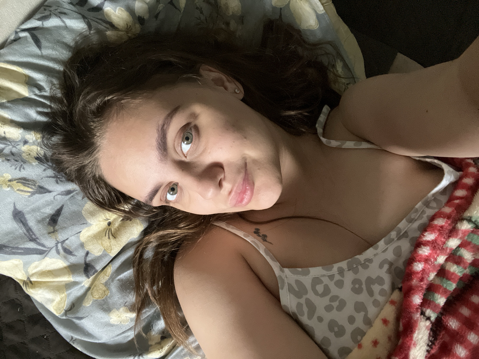A selfie shot of a fair-skinned young woman with light brown hair and green eyes, lying in bed. She is wearing a light-colored tank top with a grey animal print, partially covered by a red and white plaid blanket. A tattoo is visible on her upper chest near her shoulder. She is lying on a bed with a floral-patterned sheet. The background is dark, with hints of a dark headboard. There is no evidence to suggest a location in Chaikovsky, Russia.