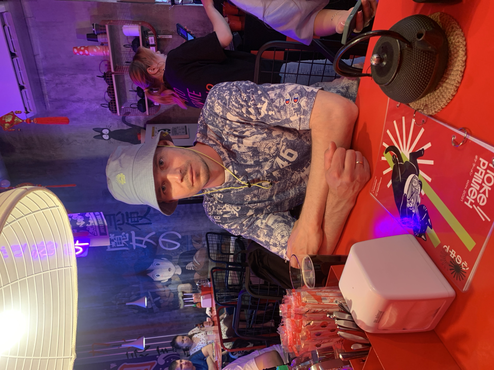 The image shows a man with a bucket hat sitting at a red table in what appears to be a restaurant in Perm, Russia. He is wearing a patterned blue shirt with the number "46" on it. His hands are clasped. On the table is a black teapot on a straw trivet, a red menu with Japanese text, a white cube (possibly a light or holder), chopsticks, and silverware. In the background, there is a wall with cartoon characters and Japanese writing. There are other people at tables in the background. The lighting is colorful, with purple and neon hues.