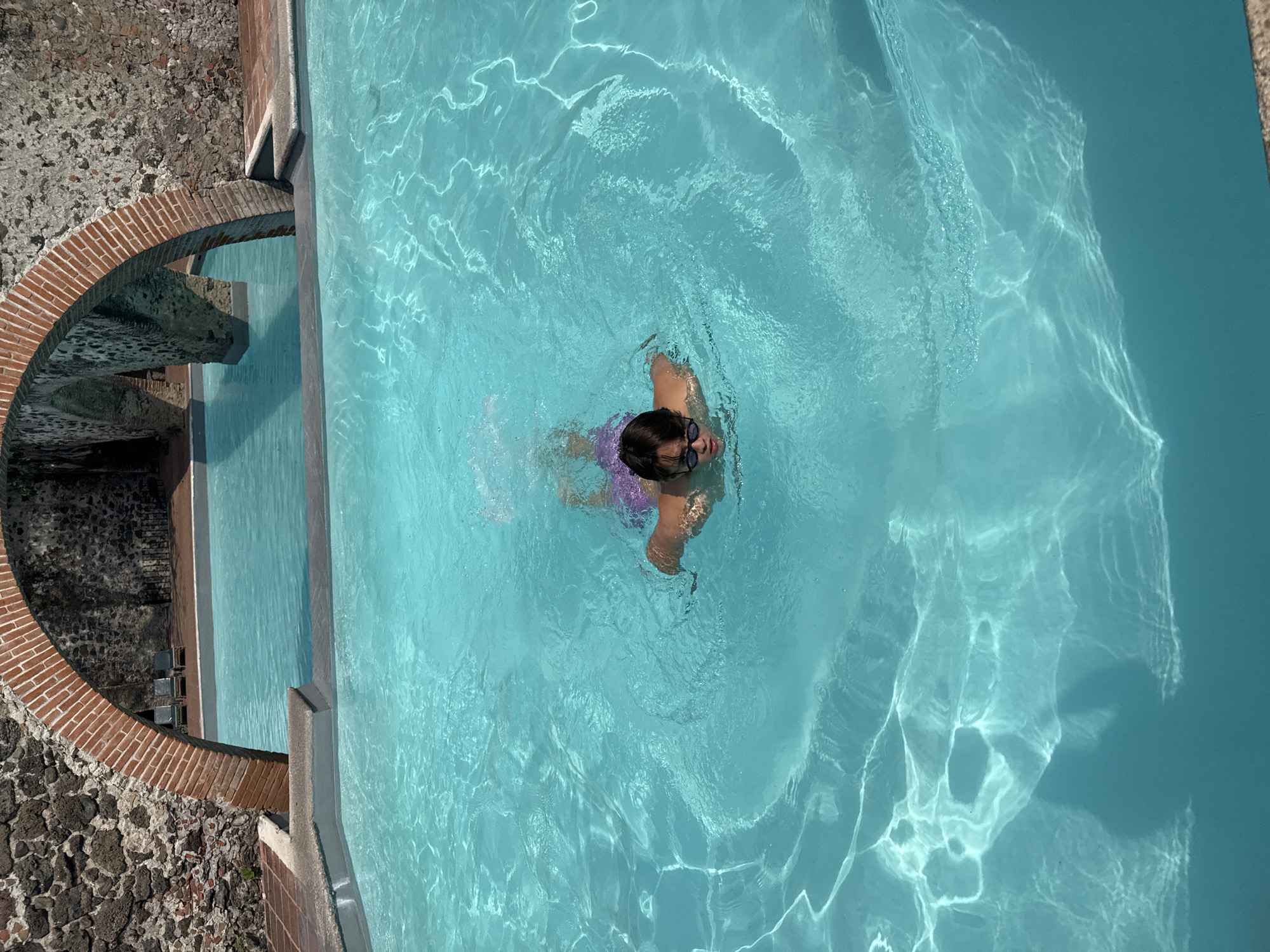 A person wearing goggles and purple swimming trunks is swimming in a blue pool. The pool is located next to a brick archway and stone wall structure. The scene is in Cocoyoc, Mexico.