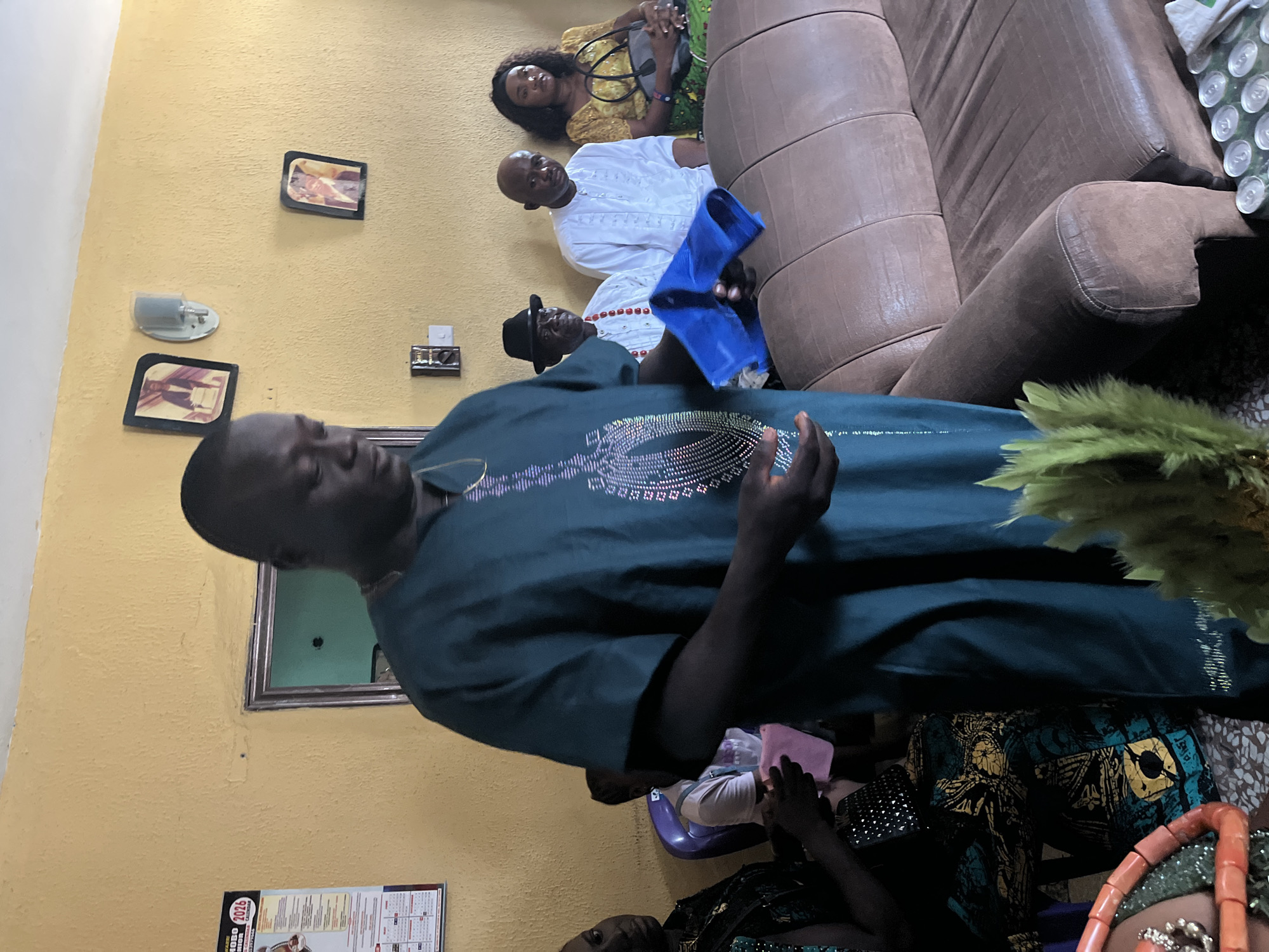The photograph appears to be taken indoors in Warri, Nigeria, within a residential setting. The room features yellow walls, adorned with framed pictures and a wall-mounted light fixture. A man in a dark green traditional attire dominates the foreground. Behind him are a few individuals: a man in a white traditional outfit, a woman in a yellow and green dress, and another person wearing a hat. A brown leather sofa occupies the right side of the frame. The lighting is indoor and possibly artificial.