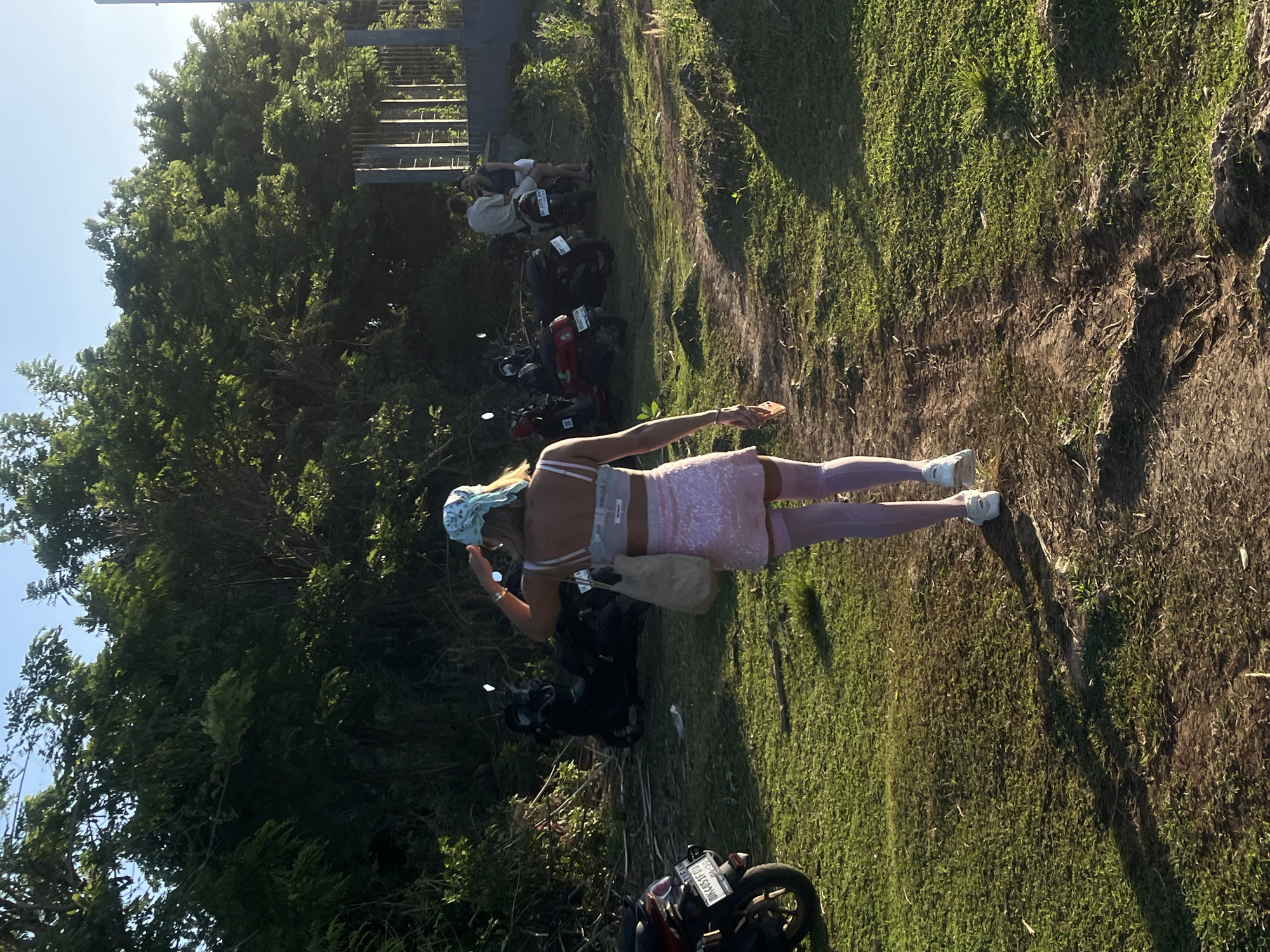 The image shows a person wearing a pink outfit and a headscarf standing on a grassy area next to a row of parked motorbikes. The location appears to be outdoors, possibly a parking area near a wooded or vegetated area. There are other people in the background, some near the motorbikes. The lighting suggests it is daytime. Based on the location of Kuta Selatan, Indonesia, there are several possible location types, including beaches, tourist sites, residential areas, or parking areas near popular attractions or accommodations. Without additional features, it is impossible to pinpoint a particular site. The person is holding an object in their hand.