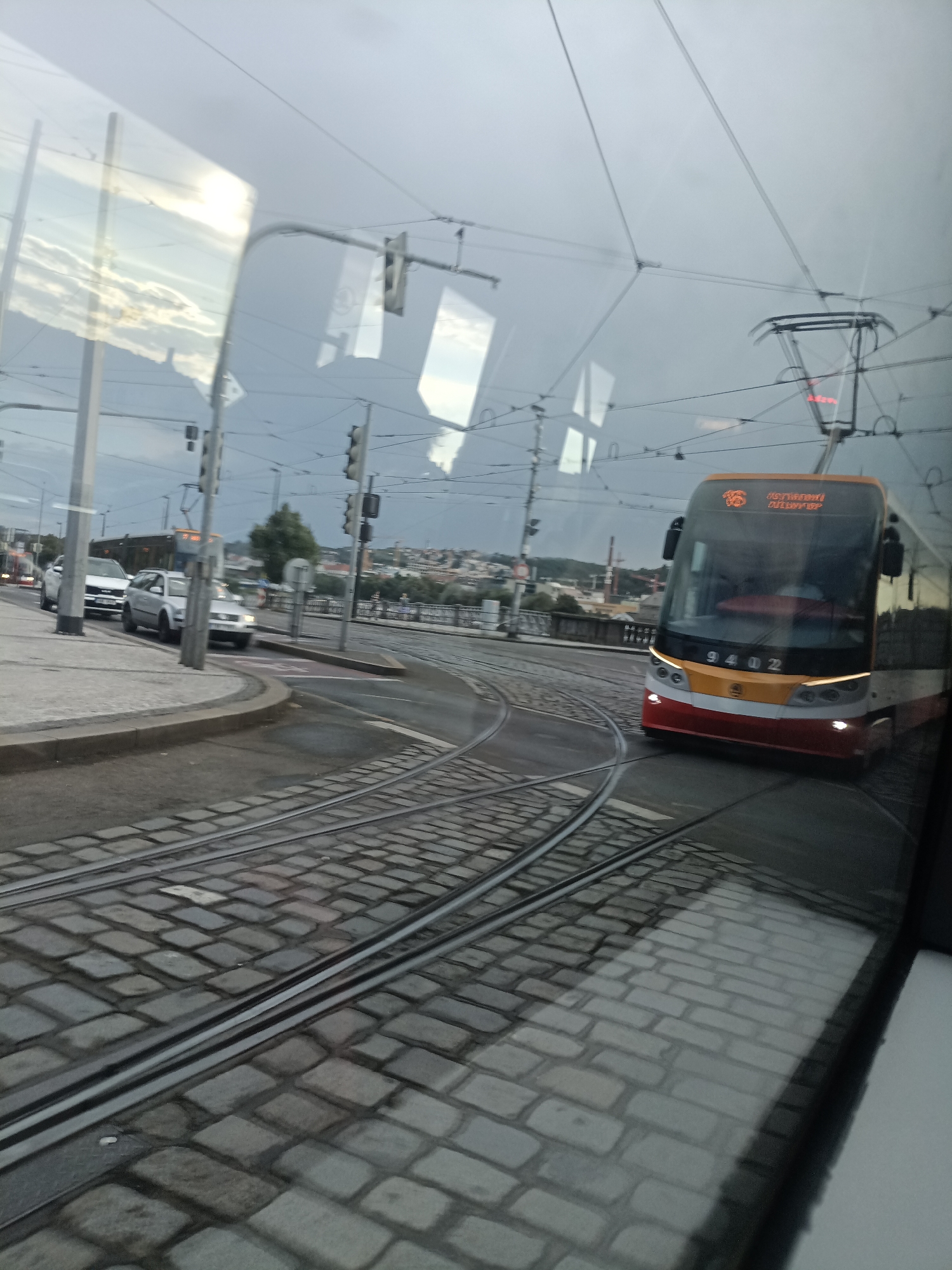 The image shows a view from a tram in Prague, Czechia. A tram is moving along a cobblestone track that curves to the right. In the foreground, the tram tracks are clearly visible. The tram itself is orange and white with the number 9402 displayed on its front. The destination display shows text in Czech, "Národní třída". To the left, there are several cars parked and moving on a paved street. In the background, buildings and trees can be seen, with a cloudy sky overhead. The reflection on the tram's window shows overhead wires and traffic lights, as well as the sky and some buildings.