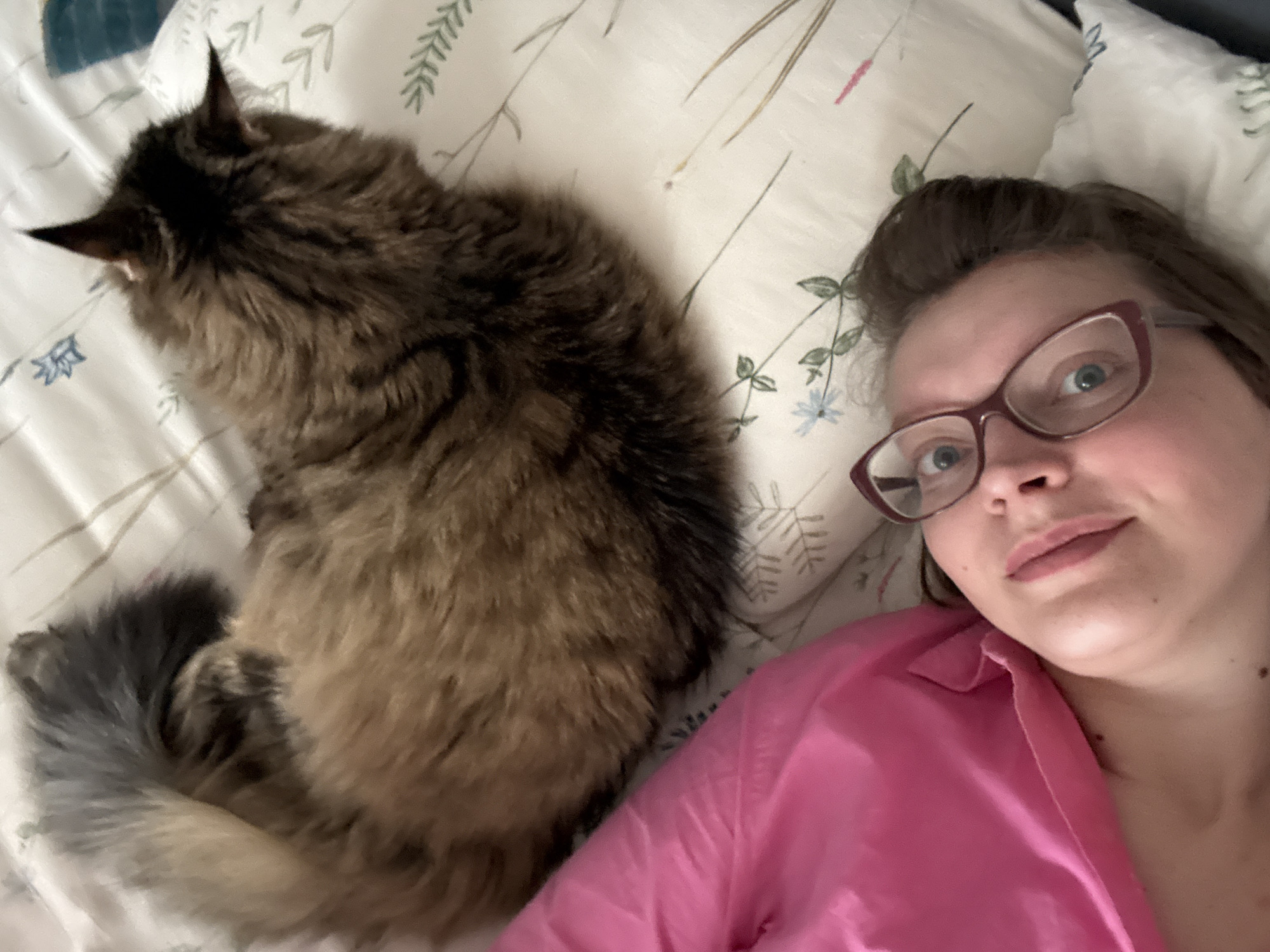 A woman and a cat are lying on a bed covered with a white floral patterned bedsheet. The woman is wearing glasses and a pink shirt, and is looking directly at the camera. The cat is brown and black, and is curled up next to the woman. The location is unknown.