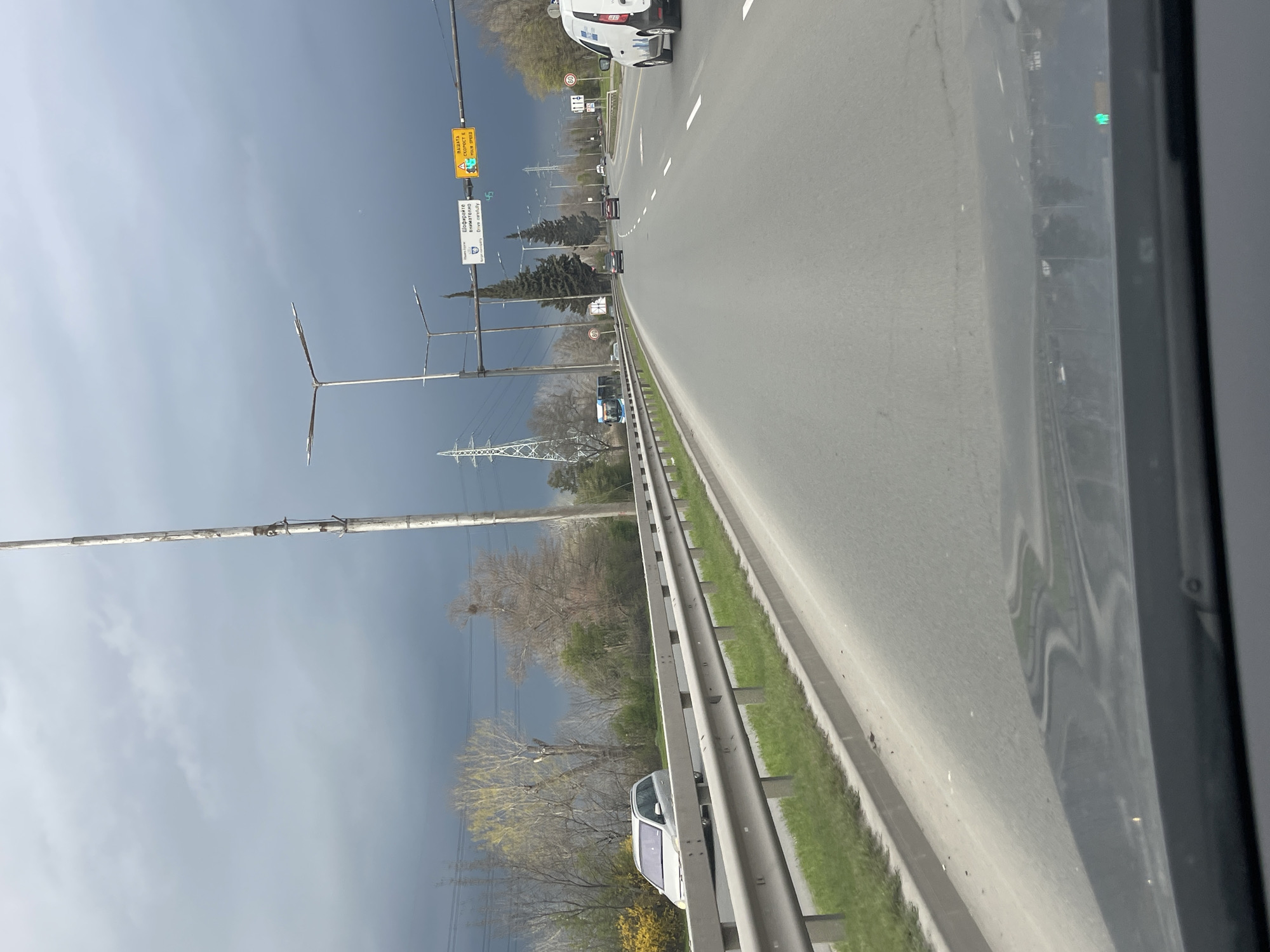 The image shows a view from inside a vehicle, looking out onto a multi-lane road. The road is paved and has dashed white lines dividing the lanes. A metal guardrail runs along the right side of the road. Beyond the guardrail, there is grass and trees. In the distance, power lines and towers are visible. The sky is overcast with dark storm clouds. There are several vehicles on the road, including a white van in the lead and other cars further ahead. Road signs are visible on the left side of the road, but their details are not clear. It is not possible to confirm the precise location in Burgas, Bulgaria, based on the visual information provided.