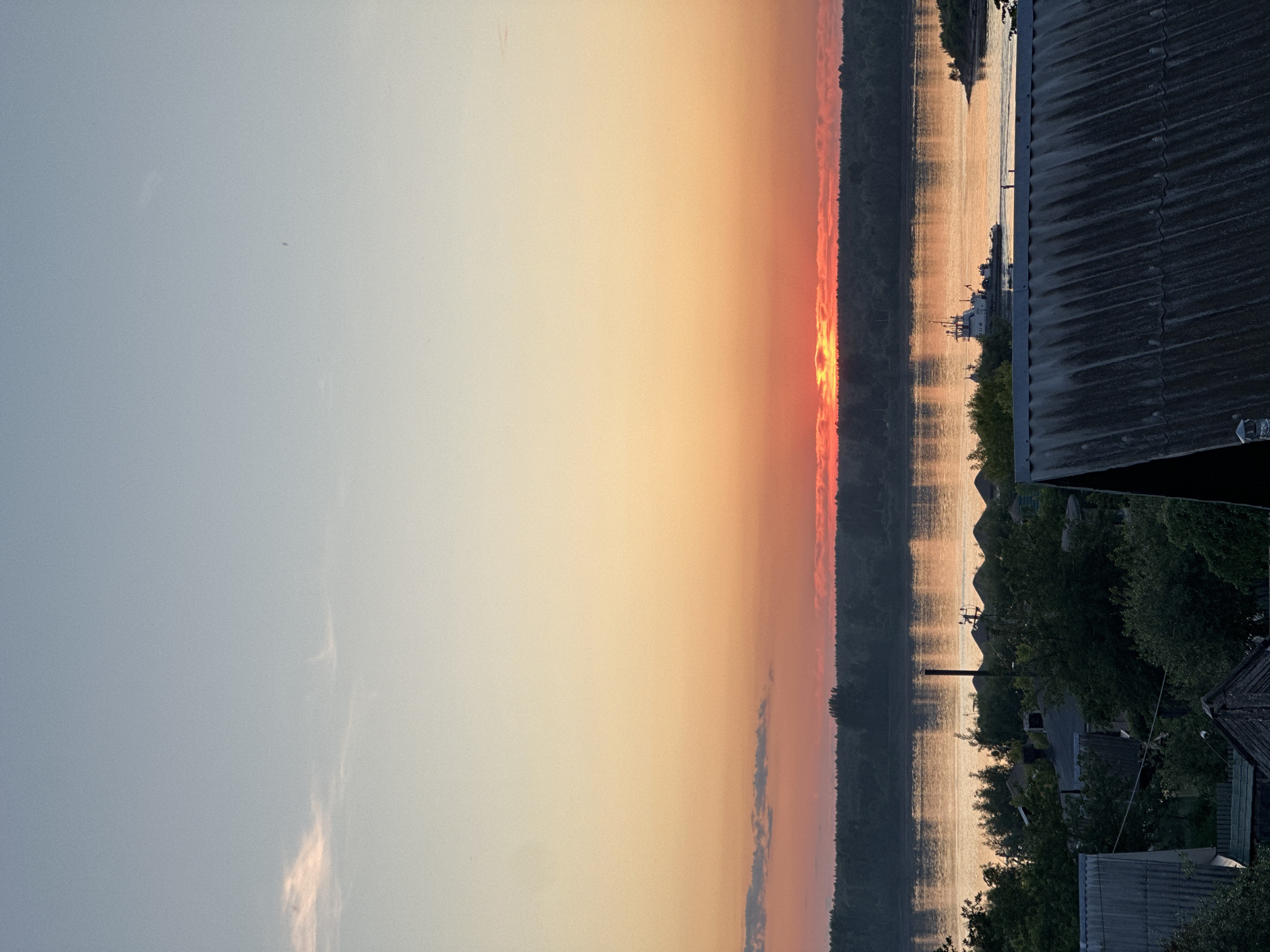 The image captures a sunset over a wide body of water, identified as likely the Kama River, within the city of Chaikovsky, Russia. The sky displays a gradient from light blue in the upper region to pale yellow and intense orange-red at the horizon. The setting sun is partially obscured by a thin band of clouds, creating a vibrant orange-yellow glow just above the distant tree line. A single, wispy cloud is discernible in the upper left portion of the sky.
A dense, dark forest defines the distant bank, appearing as a dark silhouette against the luminous horizon. The surface of the water reflects the warm hues of the sunset, exhibiting elongated, shimmering vertical streaks of light. A white and blue tugboat, bearing the partial inscription "RUB" or similar on its side, is traversing the water from left to right, generating a subtle wake. On the nearer shore in the mid-ground, several large, conical piles of light-colored material, consistent with sand or gravel, are arranged. A small, forested landmass extends into the water on the far right.
The foreground is characterized by a dense canopy of dark green trees and multiple grey roofs of buildings. A large, dark corrugated roof occupies the lower right, exhibiting visible texture and wear. Additional roofs and structural elements are partially visible through and beneath the foliage, including a tall, dark vertical pole or chimney positioned against the water's reflection in the mid-left. No individuals are depicted within the scene.