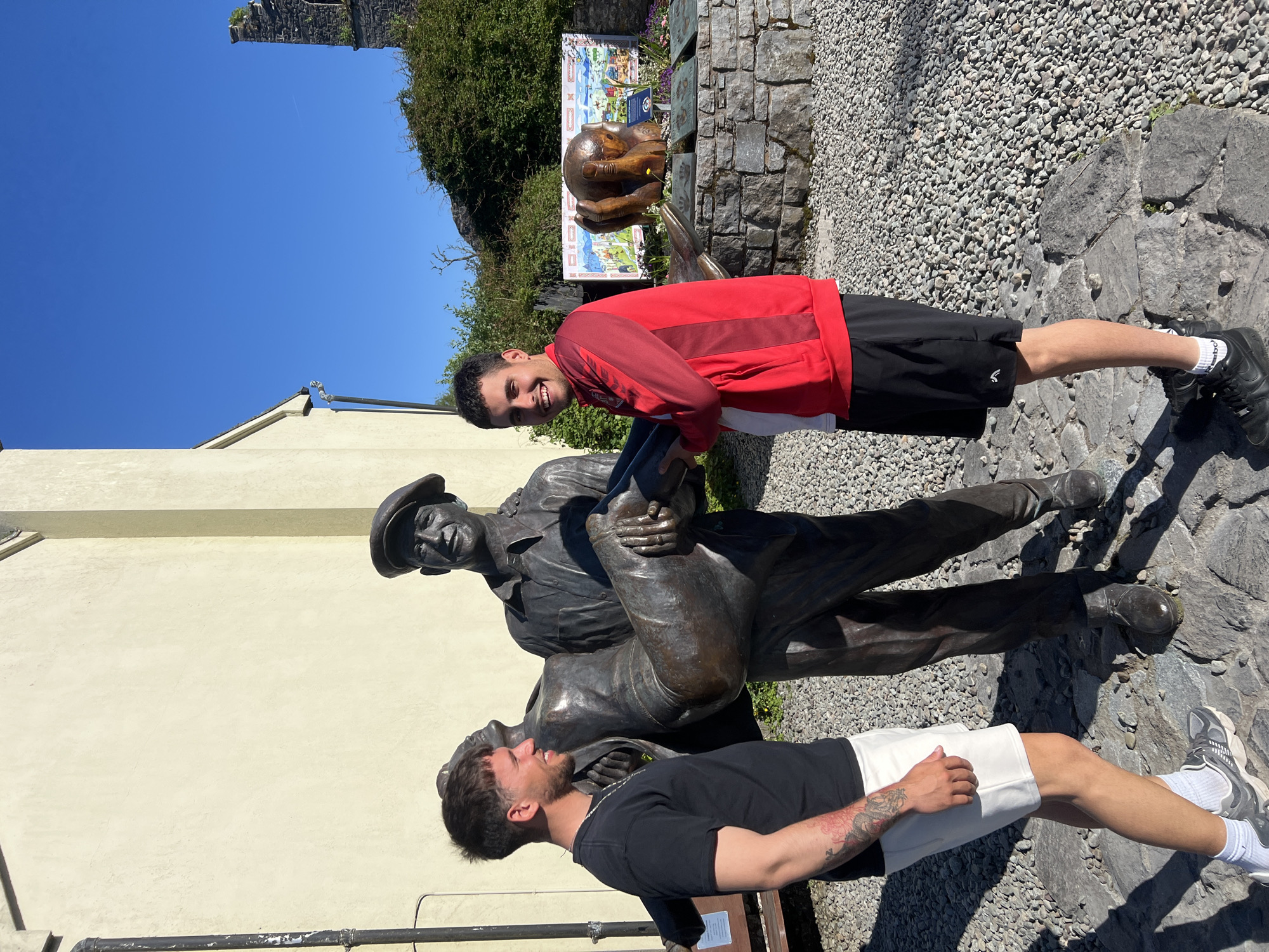 In the city of Cong, Ireland, two young men pose with a bronze statue depicting two seated figures. The first man, wearing a red long-sleeve shirt and black shorts, stands behind the statue, smiling at the camera. The second man, wearing a black t-shirt and white shorts, leans against the statue with a smile, his right arm bearing a tattoo. The statue, likely representing two seated figures, has one wearing a hat. The ground is covered with small, grey pebbles. A stone fountain is visible in the background, along with a sign displaying local attractions. The sky is clear and blue.