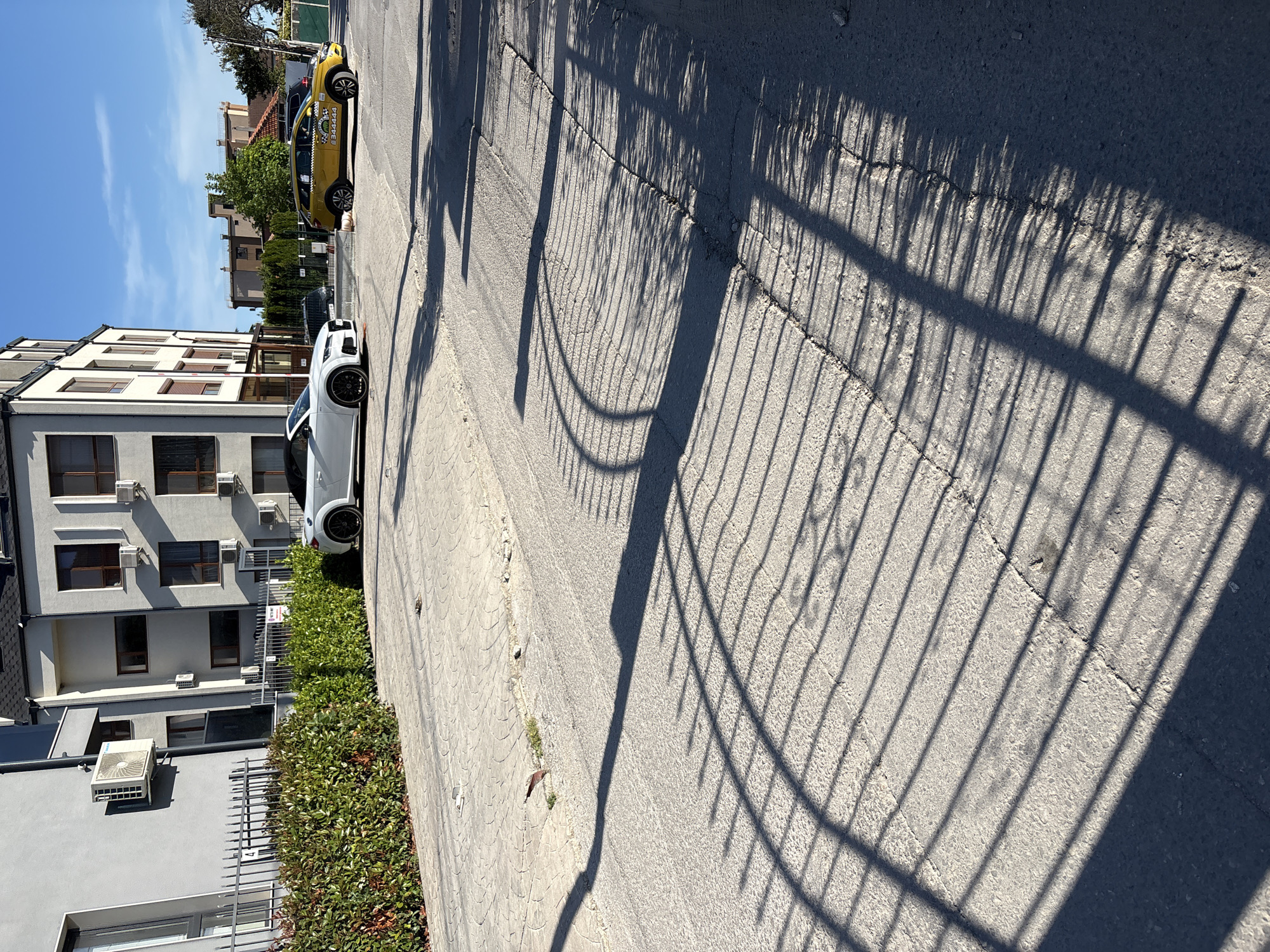 This is a daytime, eye-level, outdoor shot of a street in Varna, Bulgaria, on a sunny day. The foreground is dominated by a cracked asphalt road with prominent shadows cast by a fence. The shadows create a striped pattern across the road. The midground features a white convertible parked on the left, with a modern multi-story building in the background. To the right of the white car, a yellow taxi with a "Sunny Beach" logo is parked. The background shows more buildings and trees under a clear blue sky. The scene depicts a residential or commercial street with parked vehicles and architectural elements.