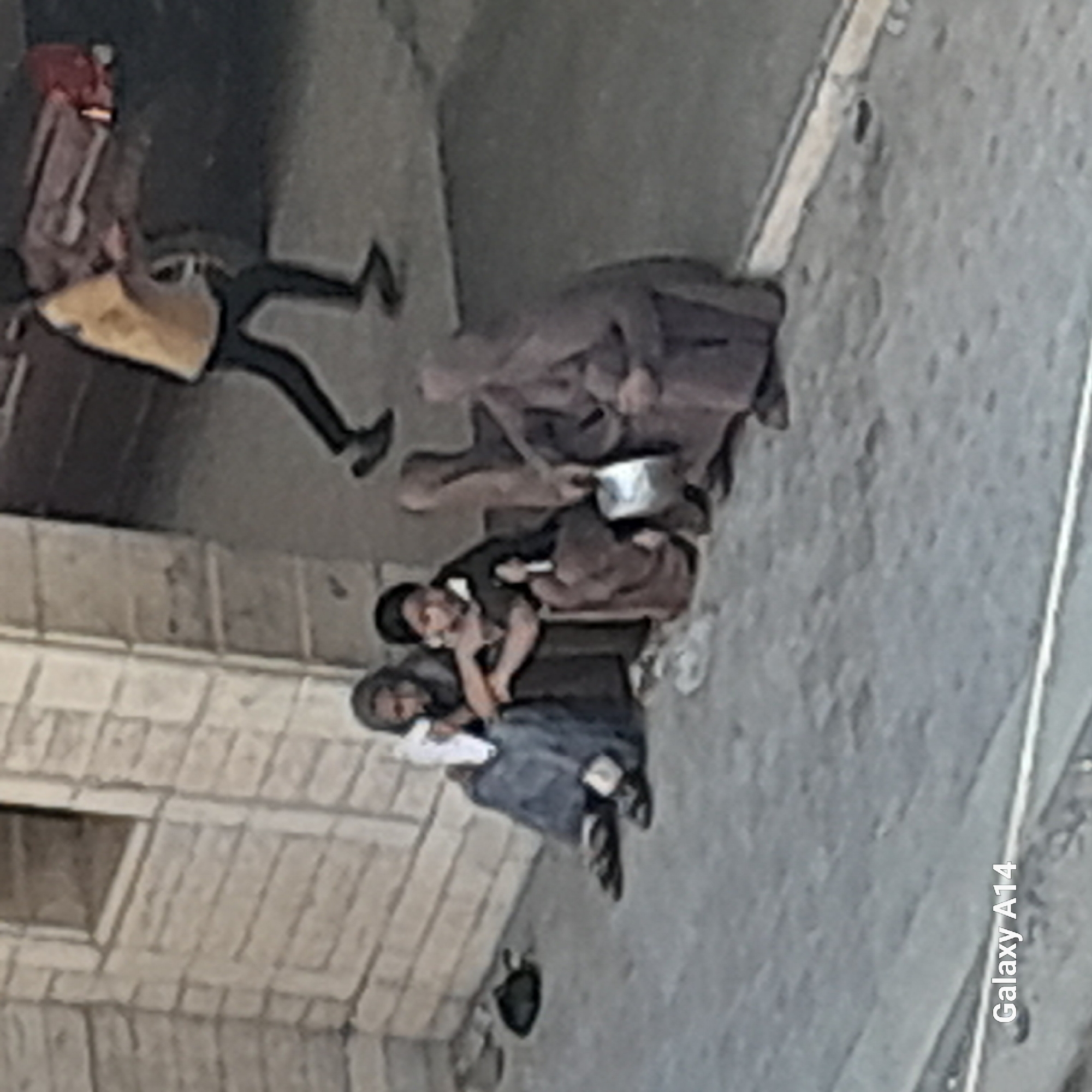 The image, taken in Khan Yunis, Palestinian Territories, depicts a street scene with several individuals. In the foreground, three people are seated on the ground. The person on the left, wearing a blue garment and a headscarf, holds a white item. Next to them, a person in a darker outfit and headscarf appears to be looking down. To their right, another individual in a brown garment is seated, holding a metal container. In the background, a person in a yellow shirt and black pants walks away, carrying a red object. The environment appears to be an urban setting with a building constructed of light-colored blocks in the background. The ground is paved with concrete. The overall impression is one of people in a public space, possibly engaged in daily activities or seeking aid.