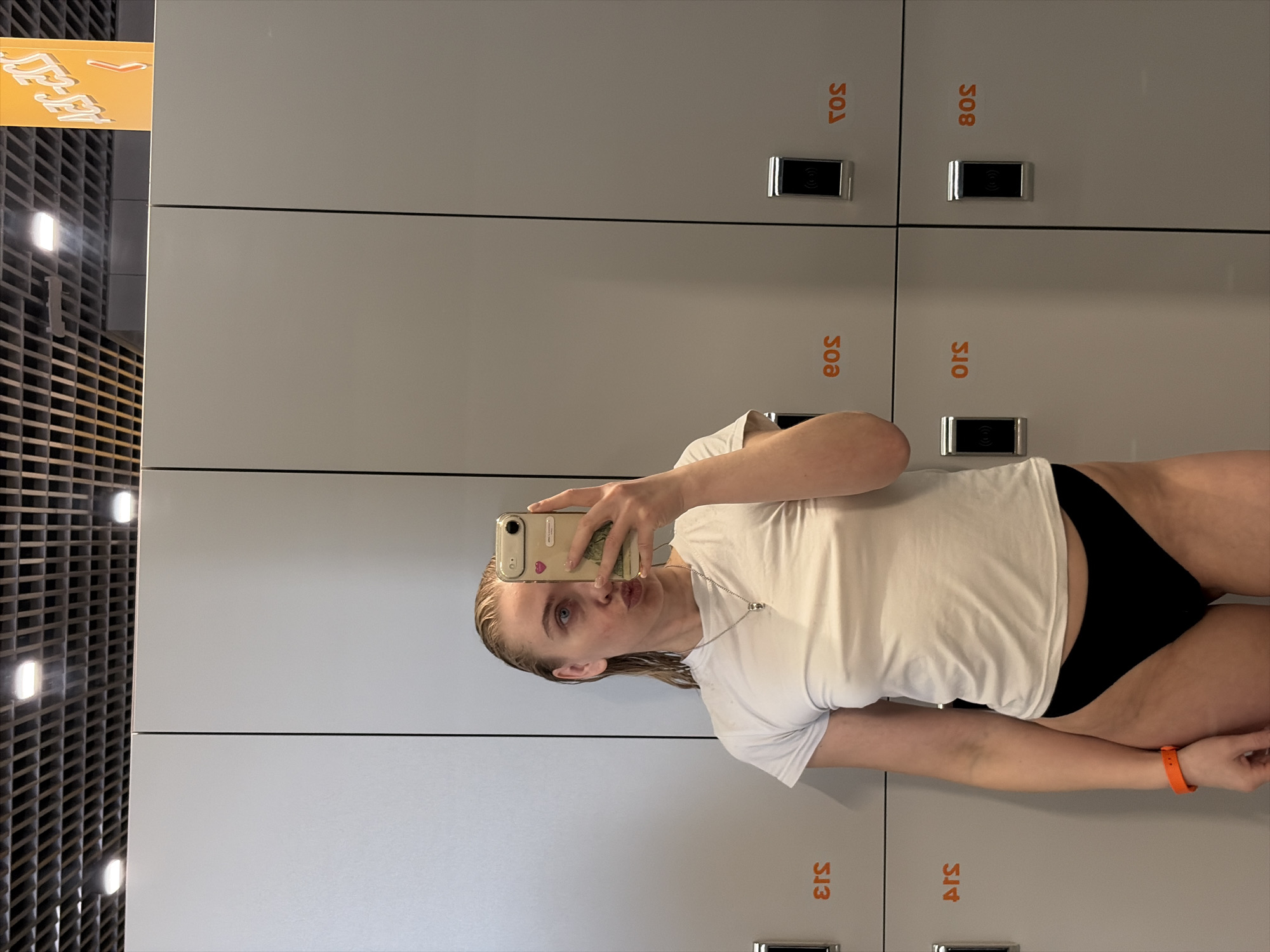A blonde-haired individual, appearing female, captures a mirror selfie in a locker room at what is identified as EOS Fitness in Votkinsk, Russia. The subject stands centrally, holding a light-colored smartphone with a decorated case to photograph her reflection. Her hair is wet, and she wears a white short-sleeved t-shirt, black bikini bottoms, a silver-colored necklace with a pendant, and an orange wristband on her left wrist. Her visible eye is blue, and her lips are pursed. The background features a wall of light gray lockers arranged in two horizontal rows. Visible locker markings, reversed in the mirror, include "S13", "EOS", "S03", "S15", and "S08" in orange text. Some lockers are equipped with dark-faced electronic locks featuring a silver trim and a wireless symbol. The ceiling above the lockers is a dark grid with integrated square light fixtures. In the upper right corner, an orange sign with white Cyrillic text "АЭЛ-ЭЭЦ" (reversed) and a left-pointing arrow is partially visible.
