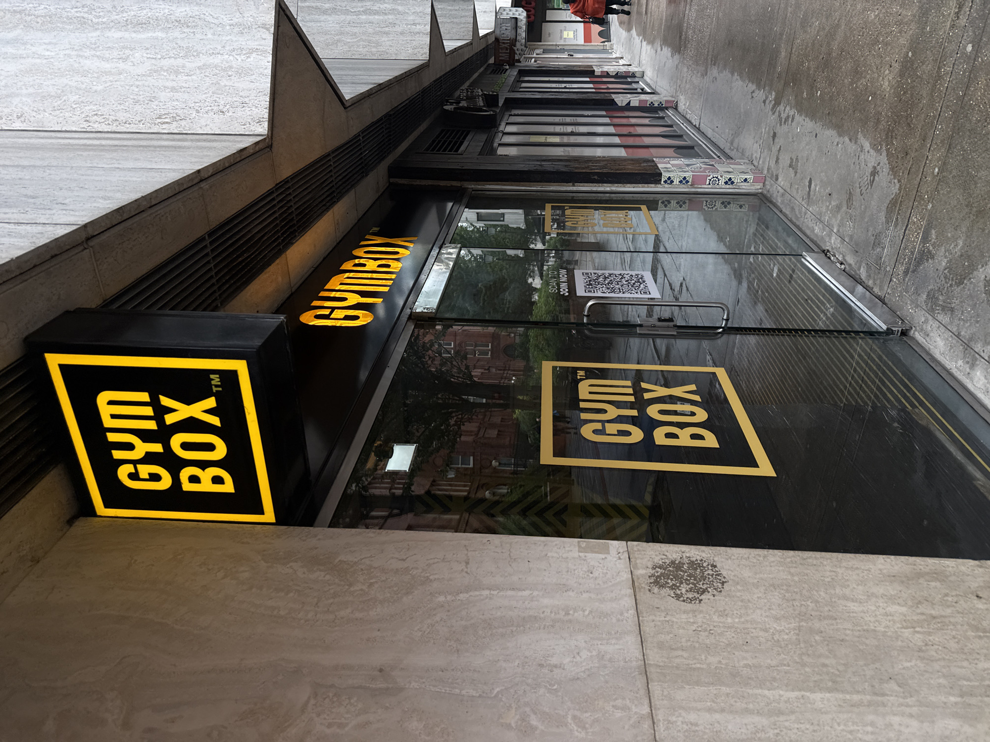 This media file captures a view of a "GYM BOX" establishment in London, United Kingdom. The scene is outdoors, with the ground appearing wet, suggesting recent rain or a damp day. The time of day is likely overcast as there are no strong shadows, and the lighting is diffused.
The primary focus is the storefront of the gym. A prominent black sign with bright yellow lettering spells out "GYM BOX" with a trademark symbol. Another similar sign is visible on the glass door of the establishment. Inside, through the glass, trees and building facades are reflected, indicating an urban environment. A QR code is visible on the glass door with the text "SCAN TO JOIN NOW". Further down the street, other businesses with signs are partially visible, including one that reads "MEXI...". In the distance, a person wearing an orange jacket is walking away from the camera.
The overall impression is of a typical street scene in a city, likely on a day with less than ideal weather conditions. The focus on the "GYM BOX" branding suggests an advertisement or a view of the business.