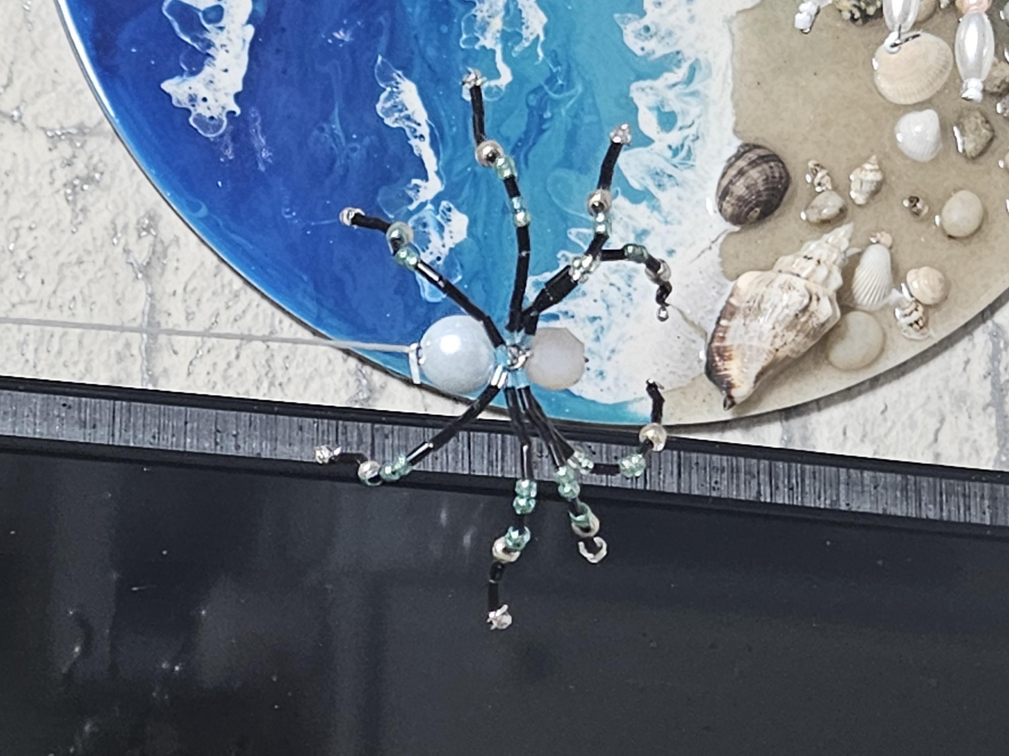 The image shows a handmade beaded spider decoration hanging in front of a dark rectangular object, possibly a phone or tablet. The spider is made of black wire and various beads, including pearl-like white beads and small green and silver beads. It has eight legs, each adorned with beads, and a larger beaded body. To the right of the spider is a circular resin art piece depicting a seascape with a sandy beach and ocean waves. The beach area of the resin art is decorated with various seashells and small stones. The background is a light-colored, textured surface, likely a table or countertop. There are no people visible in the image. The scene is indoors, and there is no information or context provided to place it specifically in the city of Kurovo, Russia.