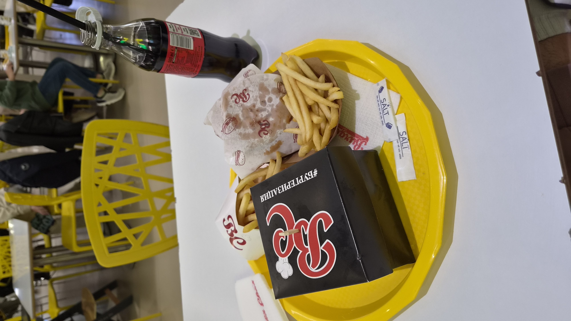 A yellow plastic tray on a white table contains a burger, french fries, a bottle of Coca-Cola with straws, and salt packets. The burger is wrapped in paper printed with "Bc" logos. The french fries are in a cardboard container and a white paper container. A black box labeled with a red "Bc" logo and the Russian word "#БУРГЕРИЗАЦИЯ" is also on the tray. A yellow chair is partially visible in the background. The image suggests a fast-food setting. There is no evidence to suggest it is located in Plovdiv, Bulgaria.