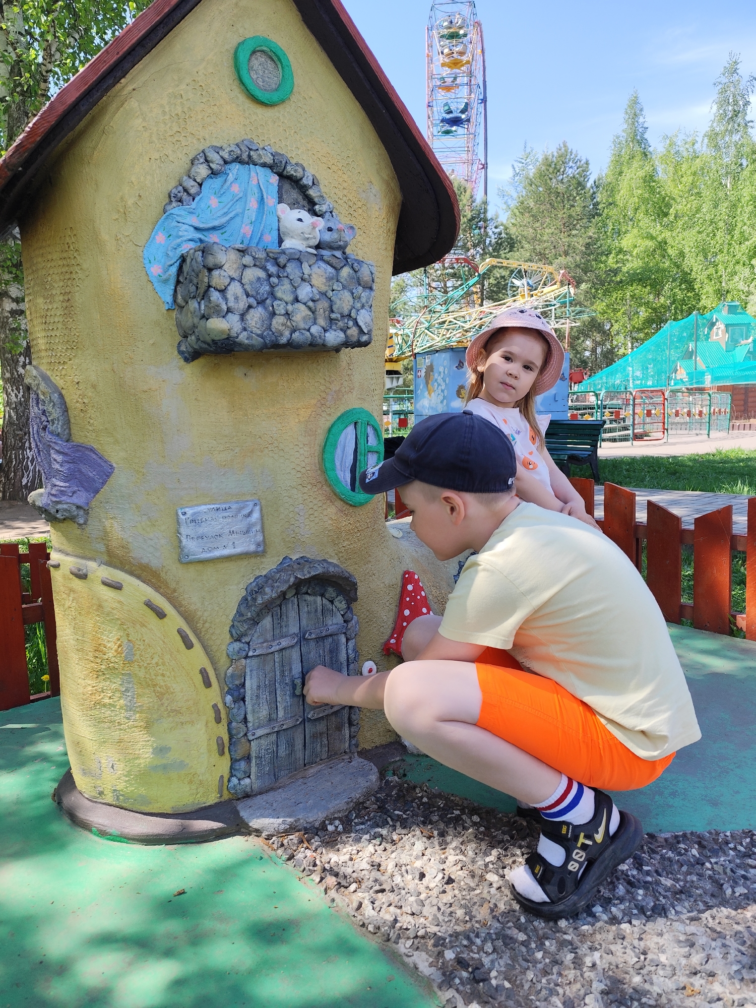 Two children are interacting with a miniature whimsical house in a public park. A boy wearing a black baseball cap, a light yellow t-shirt, and bright orange shorts is crouching down to reach the door of the house, possibly trying to open it. He is wearing white socks with colored stripes and black sandals. A girl in a pink sun hat and white shirt is standing behind him. The house itself is decorated with fairytale elements, including a balcony with mouse figurines and a small sign. In the background, there is a Ferris wheel, trees, and other park structures. The sign on the house contains Russian text, indicating the location is likely in Russia, specifically Chaikovsky.