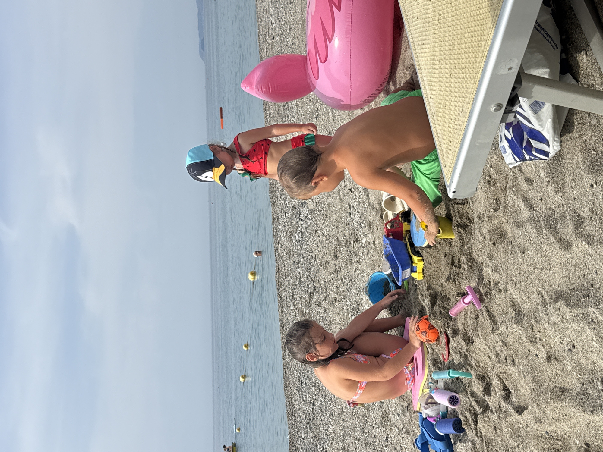 The image depicts a sunny day at a pebbly beach in Finale Ligure, Italy. The scene is populated by four young children and various beach toys. In the foreground, a young girl in a floral swimsuit is sitting on the sand, holding a pink float. Next to her, there's a collection of brightly colored toys, including a small orange ball, blue buckets, and a pink shovel. Behind her, two children are engaged in playing with sand. One boy, wearing green shorts, is partially visible, while another child in a blue and yellow penguin-themed hat is interacting with a red and green swimsuit. In the background, a large pink inflatable flamingo lies on the sand, suggesting a playful atmosphere. The sea stretches out to the horizon, with a few buoys visible in the calm water. The sky is bright and clear, indicating fair weather. The overall impression is one of carefree childhood fun on a summer day by the sea. There is visible text on a white plastic bag located under a beach chair.
