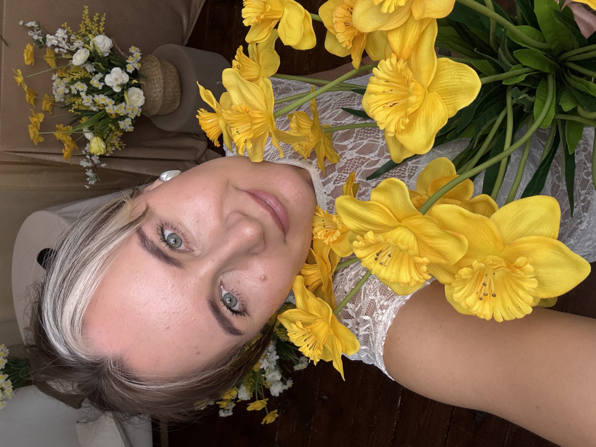 A woman with light skin, blue-green eyes, and blonde hair with a white streak is centered in the image, looking directly at the camera. She is surrounded by bright yellow daffodil flowers. She wears a white, lace-like dress. To the upper left, slightly out of focus, is a bouquet of white and yellow flowers in a woven basket. The background appears to be an interior setting with a beige object. The image was taken in Chaikovsky, Russia.