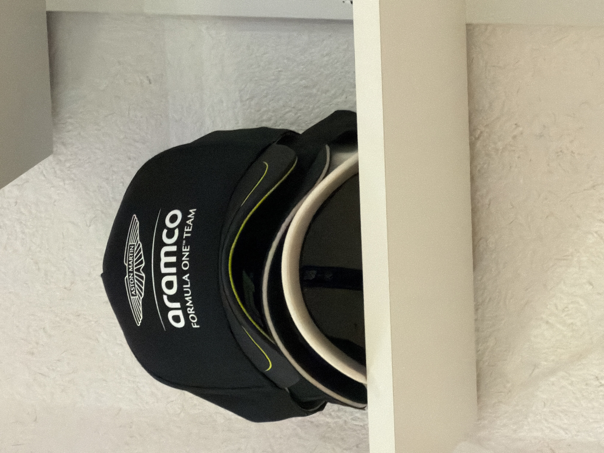 A close-up, angled shot shows a black baseball cap with the "Aston Martin Aramco Formula One Team" logo resting on a textured white surface, partially obscured by a vertical, light-colored surface that appears to be part of a drawer or cabinet. The cap features a white brim with a contrasting black band and a thin yellow accent line. The image is taken indoors, but the specific location in Kovel, Ukraine, cannot be determined from the available visual information.