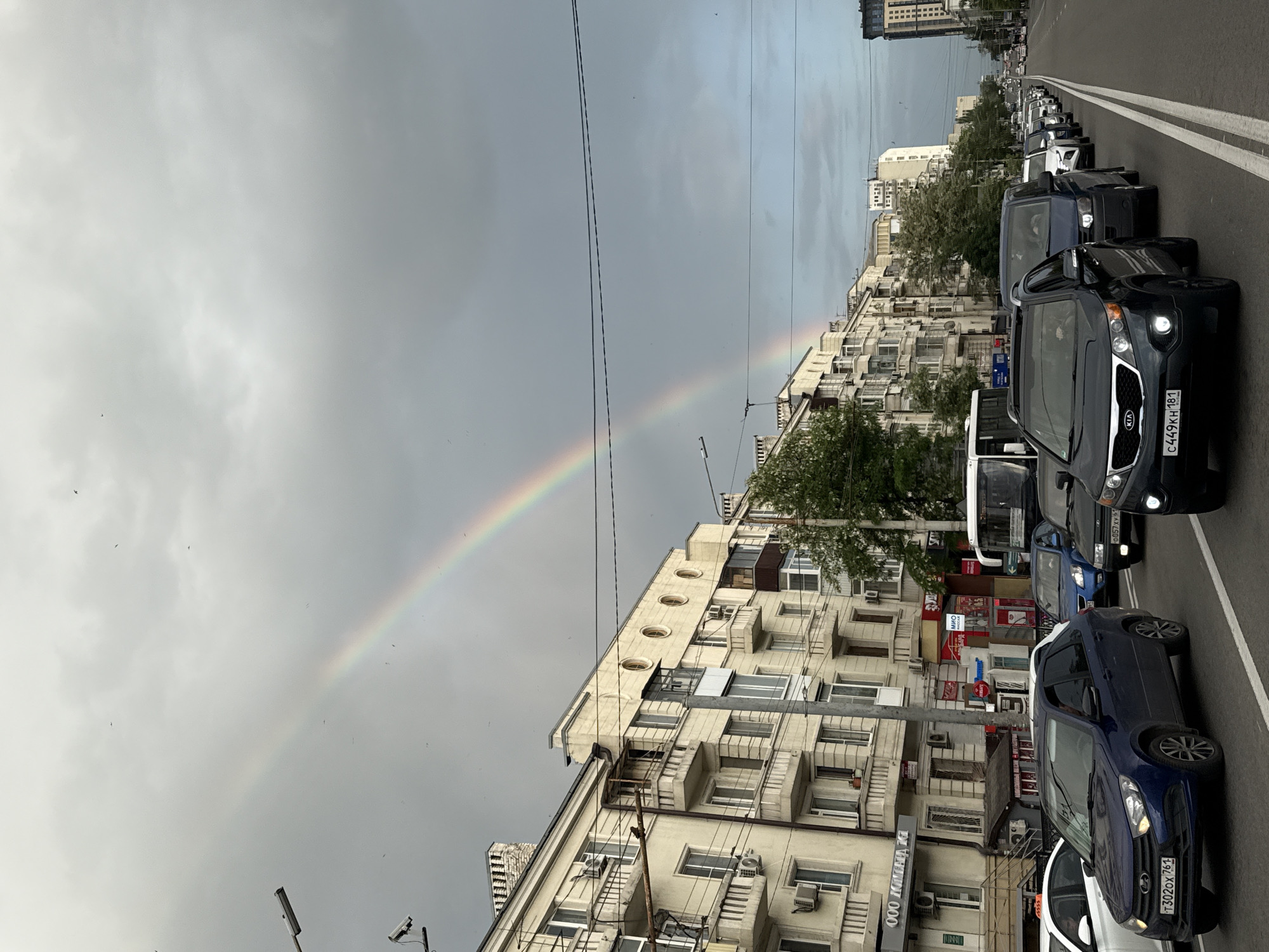 The image shows a street scene in Rostov-on-Don, Russia. A rainbow arcs across a gray sky, positioned behind a row of buildings. The buildings are light-colored, multi-story structures with repetitive window patterns and protruding air conditioning units. A line of cars is visible on the right side of the frame, creating traffic congestion on the street. Power lines stretch across the sky, intersecting the rainbow. Some birds are visible in the air. Storefront signs are visible on the buildings.