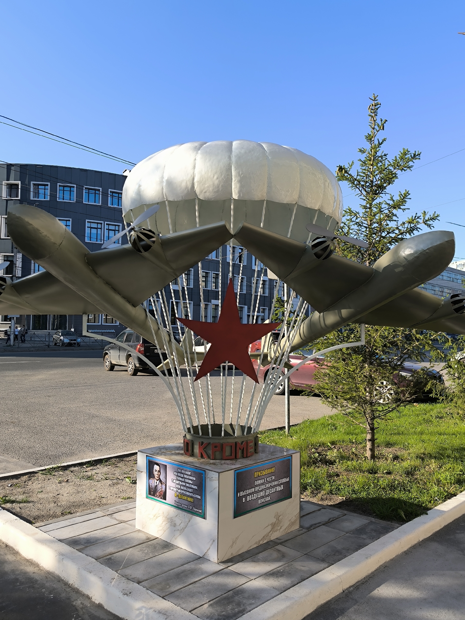 The image depicts a monument dedicated to paratroopers, featuring a large, stylized parachute with two model aircraft suspended beneath it, and a prominent red star in the center. The monument is situated outdoors in a city environment, with a modern building and a street visible in the background. Cars are parked and driving on the street, and a few pedestrians are present on the sidewalk. Green grass and a small evergreen tree surround the base of the monument. The monument has a pedestal with plaques displaying portraits and text, suggesting a historical or commemorative purpose. The clear blue sky indicates it is daytime and the weather is fair. The monument is located in Novosibirsk, Russia, specifically on the corner of Krasny Prospekt and Sibirskaya Street, near the Novosibirsk State University of Architecture and Civil Engineering.