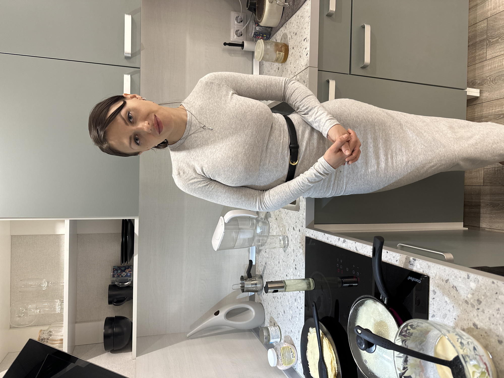 A woman is standing in a kitchen. The kitchen is located in Murino, Russia. The woman is wearing a gray dress with a black belt. She is standing near a stovetop with two pans on it, one of which has a lid. The kitchen has gray cabinets, a white shelf and light-colored countertop. The stovetop is black and smooth. There are a few bottles and kitchen utensils on the countertop. The flooring is light wood.