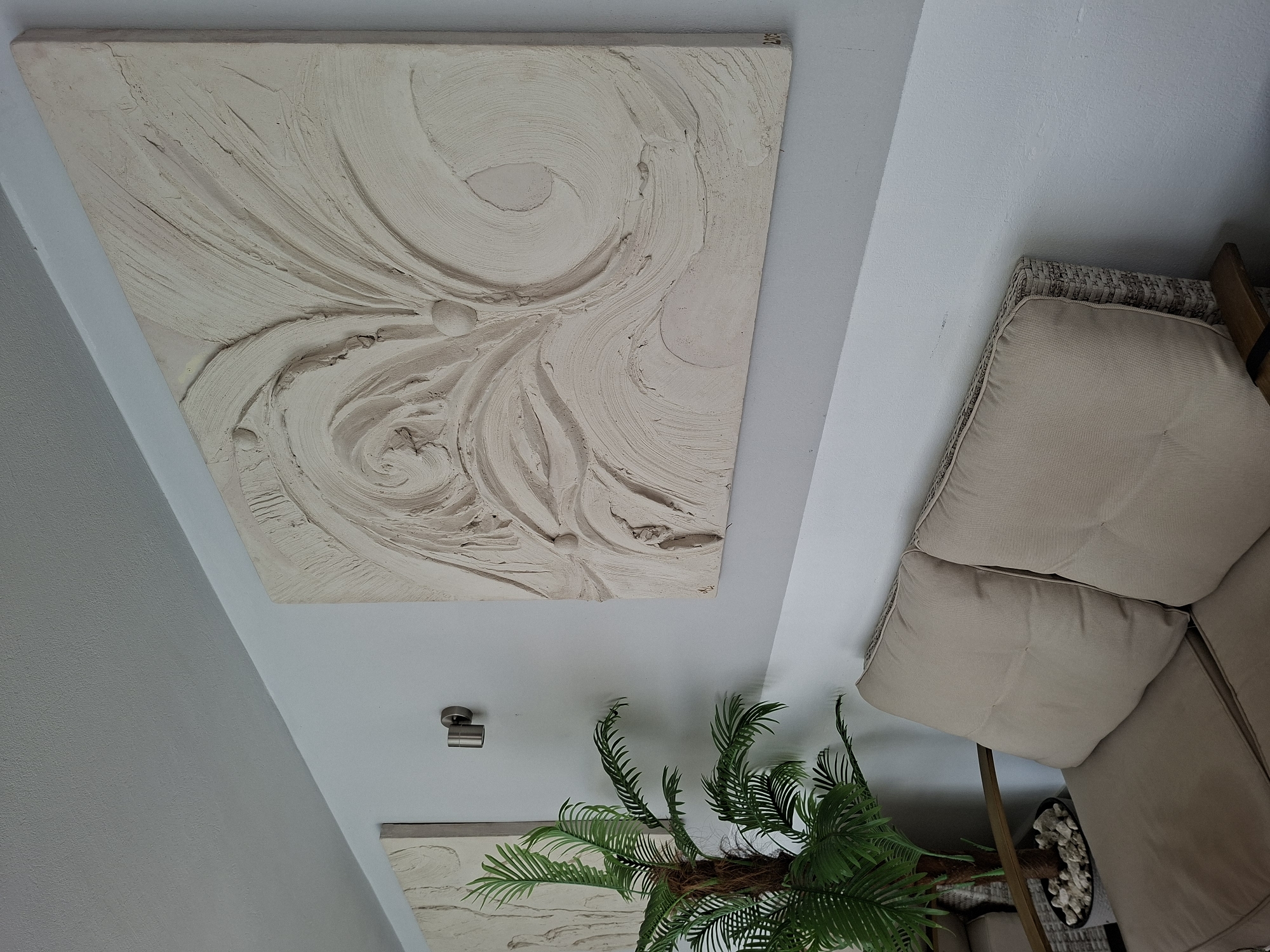 In Costești, Moldova, an indoor scene features a wall with two pieces of textured, off-white artwork mounted on it. The artwork on the right is larger and more prominently displayed, featuring swirling, organic patterns and raised spherical elements. A metal wall-mounted light fixture is positioned between the two artworks. To the left of the main artwork, a faux palm plant in a grey pot filled with white stones stands beside a beige upholstered sofa. The sofa is made of wicker and has multiple cushions. The wall and ceiling are painted in a light gray hue.