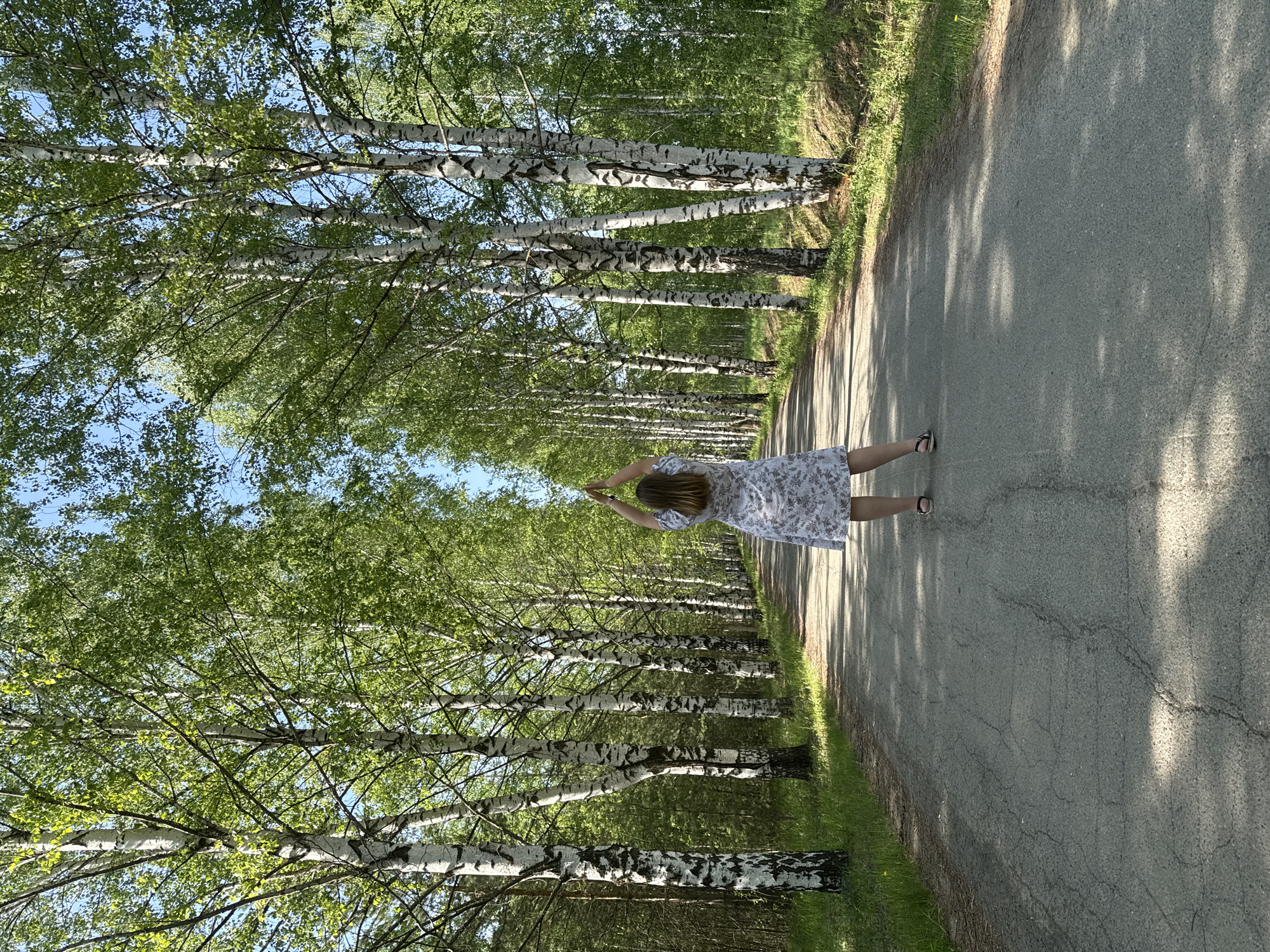 A medium shot depicts a person standing on a gray asphalt road in Банное, Russia, surrounded by trees. The person, wearing a light-colored, floral-patterned dress and dark sandals, has their arms raised above their head. The road is flanked by trees on both sides, casting shadows on the pavement. The environment consists of a forest with birch trees and green foliage. The sky is visible through the trees.