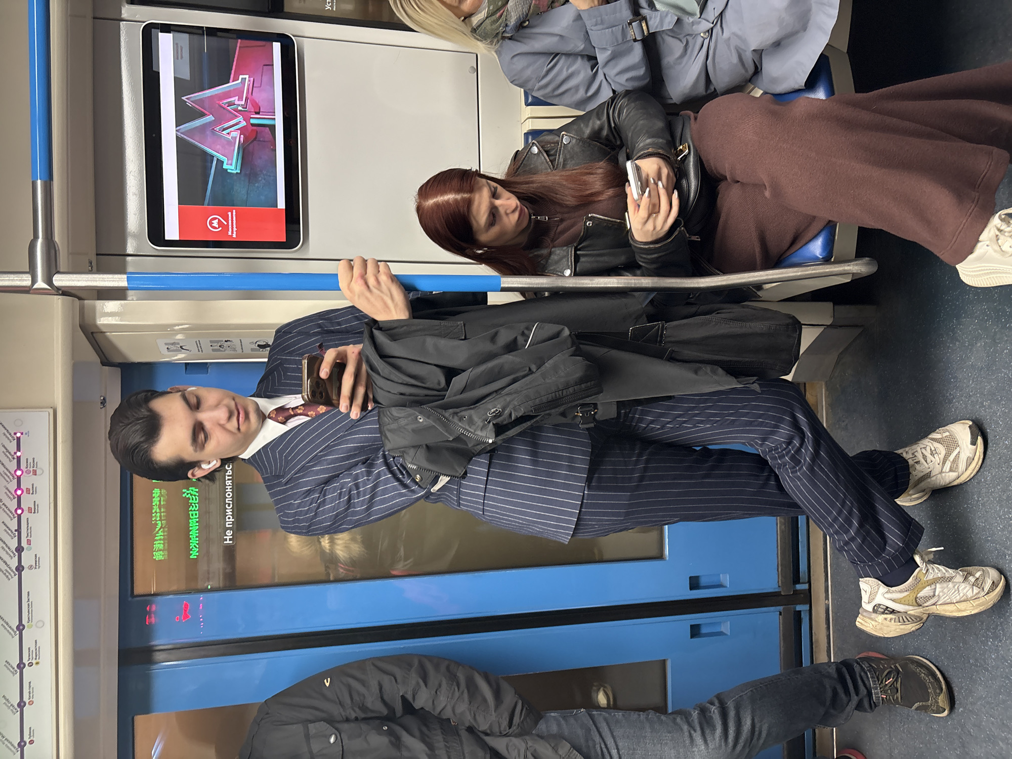 Inside a Moscow, Russia, subway car, a man in a pinstripe suit stands holding a black coat, looking at his mobile phone. He wears white wireless earbuds and white and tan sneakers. A woman with auburn hair and a black leather jacket sits next to him, also looking at her mobile phone. An older woman wearing a gray jacket is seen in the corner. A subway map is visible on the left, and a digital screen displays the Moscow Metro logo. A sign on the subway car door reads, "Не прислоняться," which translates to "Do not lean." Another person in a black jacket is visible standing in the front.