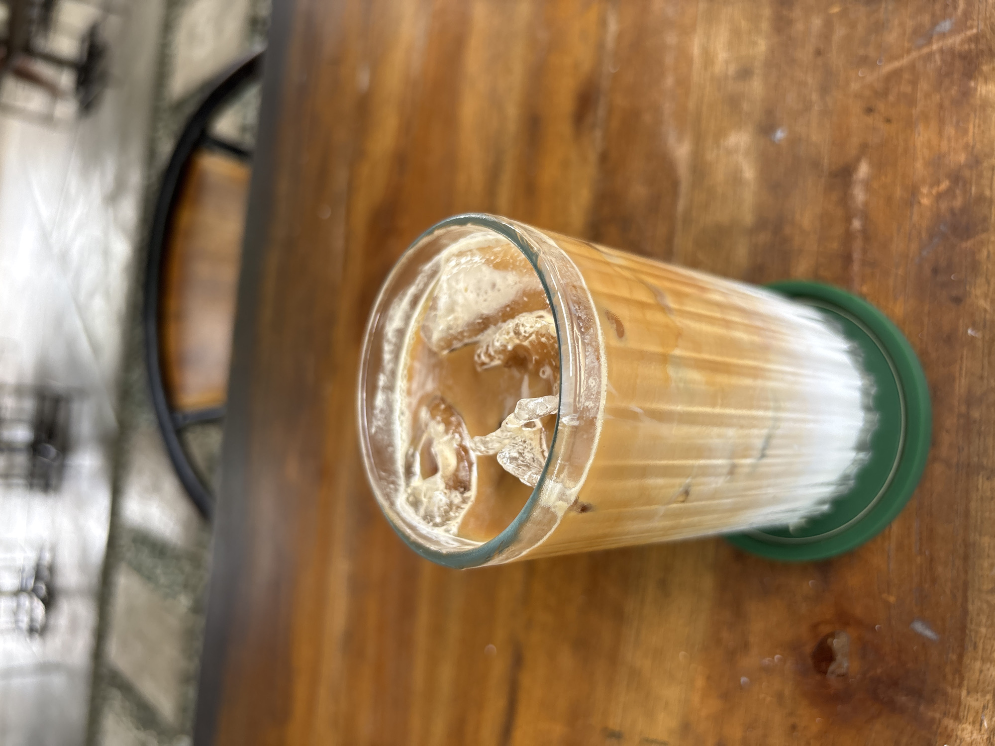 The image shows an iced coffee in a glass with a green coaster on a wooden table. The coffee appears to be layered, with a light-colored base (likely milk or creamer) and a darker coffee layer above. Ice cubes are visible in the upper part of the glass. The table has a visible grain pattern. The location is possibly in Kuta Selatan, Indonesia, given the prompt's request. A chair is also present in the background, blurred.