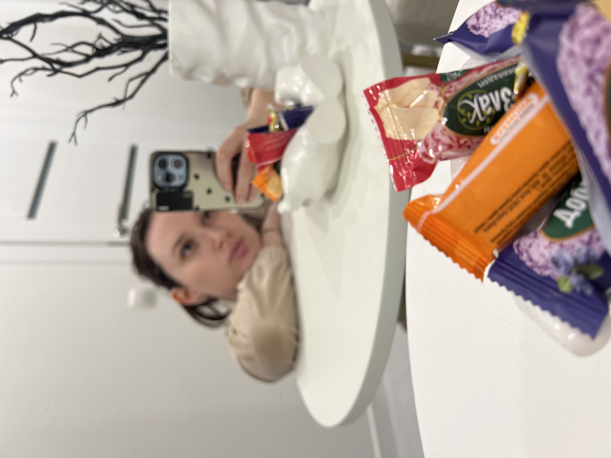 This media file captures a mirror selfie taken by a young woman in an indoor setting, likely in Tyumen, Russia. The original image is rotated 90 degrees clockwise.
In the reflection, a woman with dark hair, wearing a light-colored top, is visible. She is holding a smartphone with a patterned case, seemingly using it to take the picture. Her expression is neutral as she looks towards the phone.
The reflection also shows a white, possibly round, surface on which several packaged snacks or candies are placed. Among these are a red package with green branding, showing Cyrillic text that appears to be "Хлеб Деревенский" (Rustic Bread) and the brand "Эллада". There is also an orange package, partially visible with "ОПТИМА" (Optima) text, and two distinct purple packages. One purple package clearly displays "Долька КЛУБНИКА" (Strawberry Slice/Segment), indicating strawberry-flavored candy, while the other shows "БРУСНИКА" (Lingonberry).
Behind the white surface, in the upper part of the reflection, black, branch-like decorative elements are visible, suggesting wall art or a decorative plant. The overall setting appears to be a brightly lit, clean indoor space, possibly a home. The activity depicted is a candid mirror selfie, capturing a casual moment.