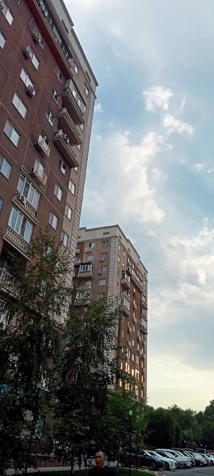 The image depicts a city scene in Almaty, Kazakhstan, featuring two apartment buildings against a cloudy sky. The building on the left is tall and red-brown, with numerous windows, balconies, and air conditioning units. A dense canopy of green trees partially obscures the lower floors of this building. The building on the right is lighter in color and appears to be of similar height, also featuring many windows and balconies. The setting sun reflects off some of the windows, creating streaks of light. In the foreground, a line of parked cars is visible, and a single man stands looking towards the right side of the frame. The overall impression is a residential urban environment, likely in the late afternoon or early evening.