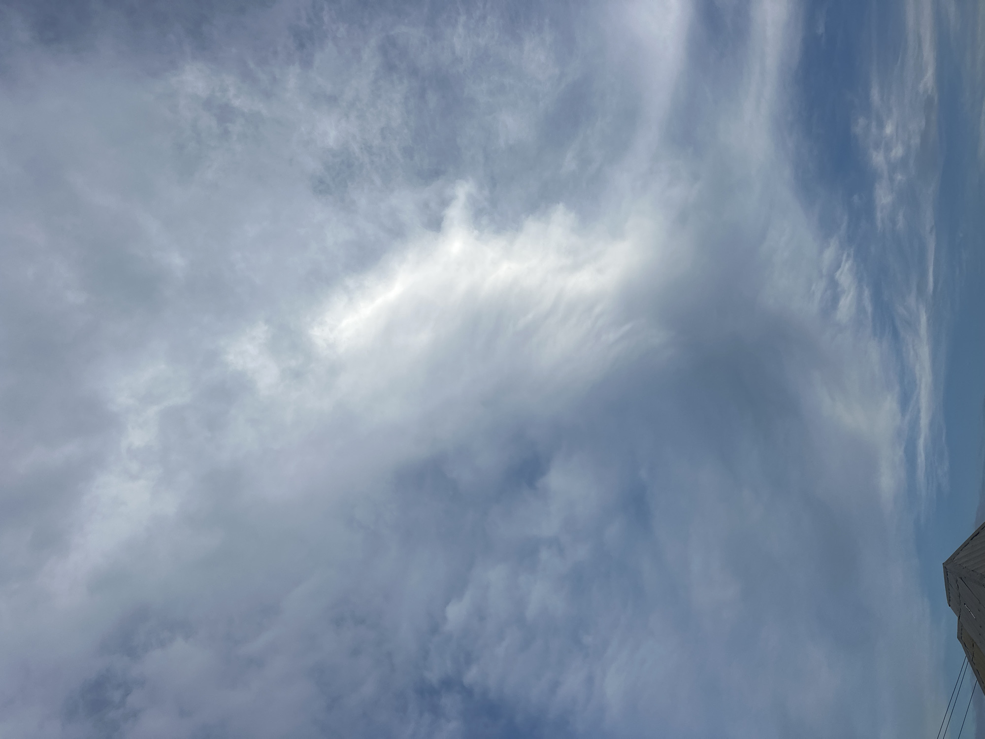 The image displays a cloudy sky with the top of a building's roof visible at the bottom left. The sky is filled with various cloud formations, ranging from thin, wispy cirrus clouds to denser, more opaque clouds, creating a dynamic and textured canopy. A bright streak of light pierces through the clouds in the upper center of the image, suggesting the presence of the sun behind the overcast. The building's roof appears to be made of corrugated metal, with a geometric, angular design. The overall lighting suggests it is daytime, but the heavy cloud cover creates a somewhat muted and diffused light. The scene is likely an outdoor shot, possibly capturing a moment before or after rainfall, given the nature of the clouds. The location is stated to be Kurunduma, Nigeria.