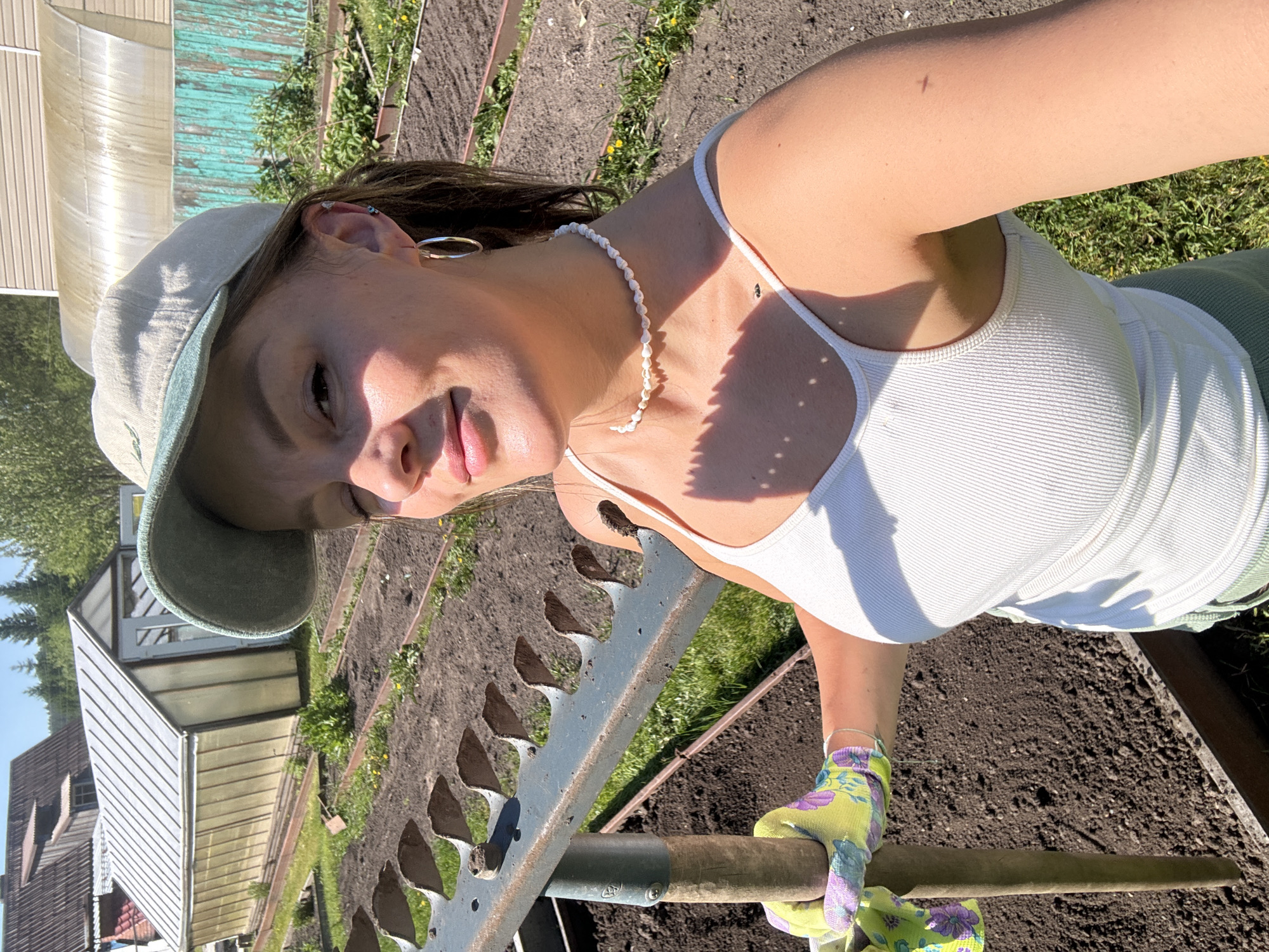 A female subject, appearing to be in her 20s or 30s, is depicted taking a selfie in an outdoor garden setting in Malaya Kuskunka, Russia. She is wearing a light-colored baseball cap with a green brim, a white ribbed tank top, and light green shorts or pants. Her left hand, clad in a yellow and purple floral-patterned gardening glove, grasps the wooden handle of a large metal rake. She wears silver hoop earrings and a pearl choker necklace. The background features several prepared garden beds with tilled dark soil, interspersed with grass and scattered dandelions. Two greenhouses are visible: a rectangular one with a white door and translucent panels, and a larger hoop-style greenhouse further back. Beyond these are a wooden house with a dark roof and a light-sided building, separated by a worn turquoise wooden fence. The scene is illuminated by strong sunlight, casting shadows.