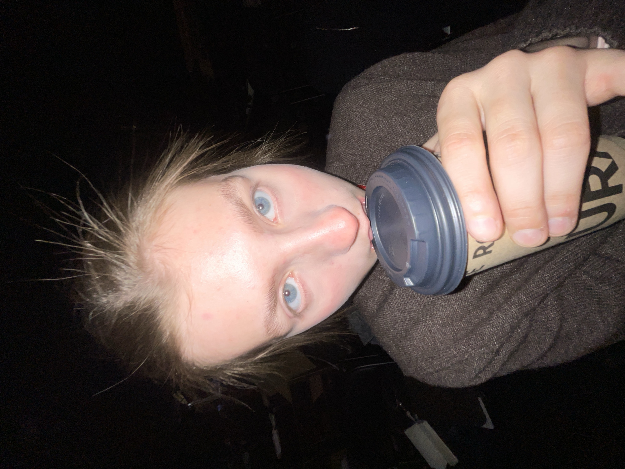 A person with fair skin and light blue eyes is drinking from a paper cup with a dark gray plastic lid. The person is wearing a brown garment. The background is dark. The location cannot be determined based on the image.