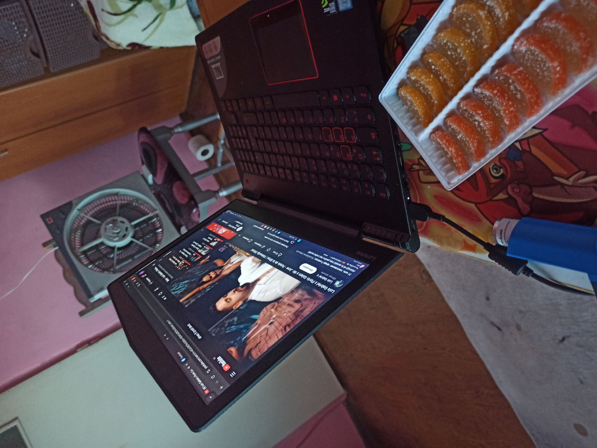 A tilted overhead shot shows a black laptop with a red trim and keyboard, partially open and displaying a YouTube video by "Luis Gabriel x Florin Salam x Mr. Juve - Fetele de la Birou." Next to the laptop sits a rectangular plastic container filled with candied fruit slices, orange and red in color. A blue and black device with a cord is also visible on the right side of the frame. Other items in the background include a fan and a set of wicker baskets atop a wooden dresser. The image's location and specific details of the city of Unknown, Unknown cannot be determined from the scene.