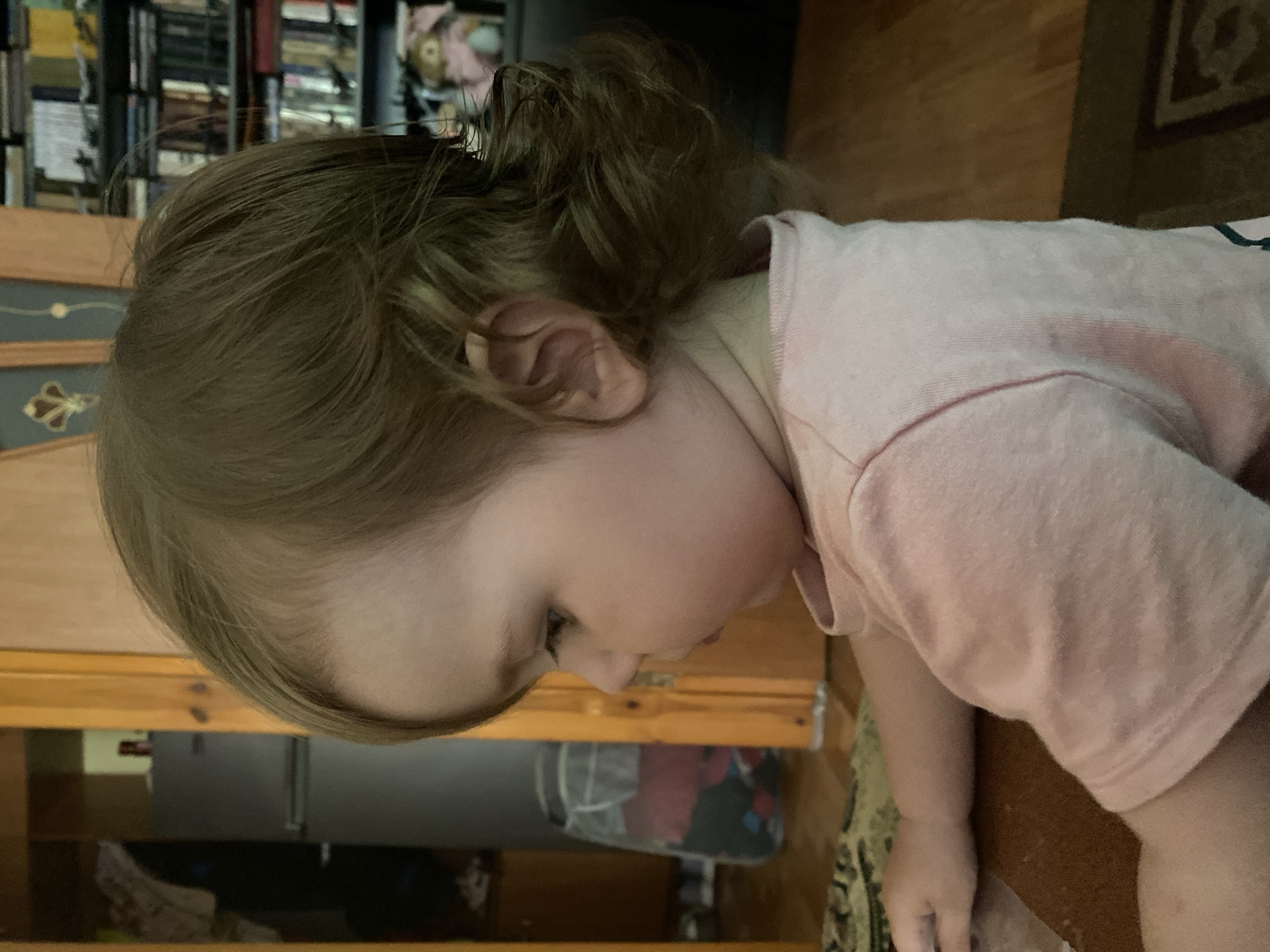 A side profile close-up of a young child, seen from the chest up, wearing a pale pink t-shirt. The child has light brown hair styled to the side and is looking down and to the left. The background is blurred, with a wooden doorframe and a glimpse of a refrigerator to the left, and bookshelves filled with books to the right. The scene is indoors, with the lighting suggesting it is daytime.