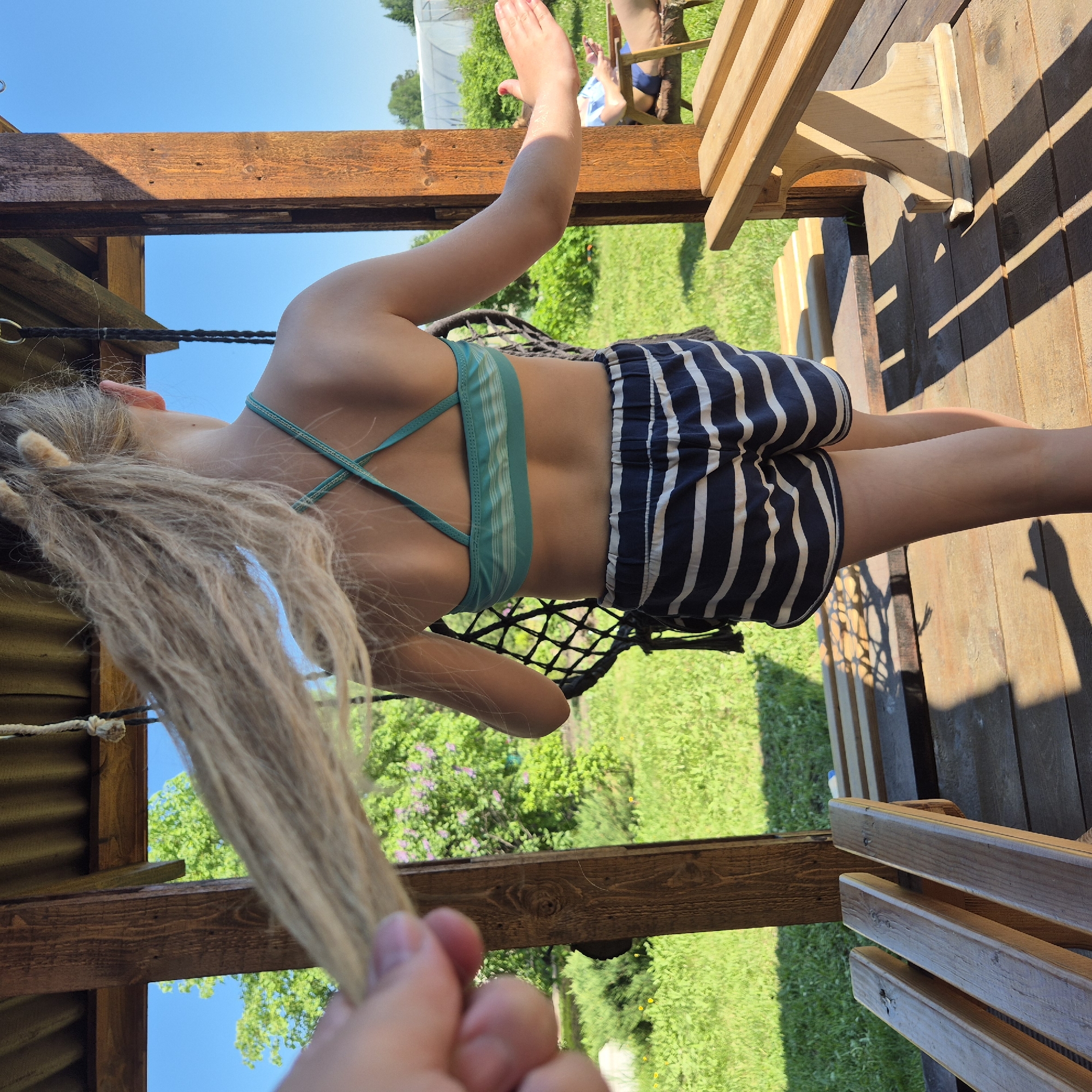The image depicts an outdoor scene in Novostroyka, Russia, on a bright, sunny day. The primary subject is a person, observed from behind, standing on a wooden deck within a shaded wooden structure, possibly a gazebo or pergola. This individual has light-colored hair, tied in a high ponytail, a portion of which is being held by a disembodied hand visible in the immediate foreground. The person wears a teal-colored sports bra or bikini top with horizontal stripes and criss-cross straps on the back, paired with navy blue and white striped shorts. The right arm is extended outwards, while the left arm is positioned near a black, net-like object, possibly a swing or hammock component, hanging from the wooden structure.
To the right of the primary subject, a light-colored wooden bench rests on the deck. Further in the background, another individual is partially visible, seated in a wooden chair, holding what appears to be a smartphone or small device. This person is wearing light blue attire. Beyond the wooden structure, the environment consists of vibrant green grass, various trees, and bushes, with some purple flowering plants visible in the mid-ground. A clear blue sky dominates the background, and a white, translucent greenhouse or hoop house structure is discernible in the far distance on the right. Strong sunlight casts distinct shadows of the wooden structure's beams onto the deck.