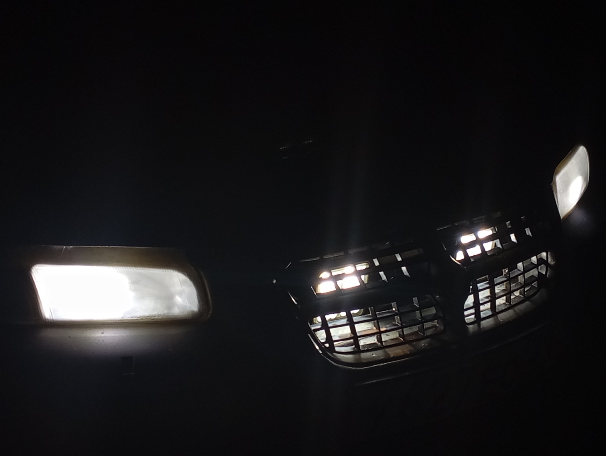The image depicts the front fascia of a dark-colored vehicle at night in Kurganinsk, Russia, illuminated by its own lighting. On the left, a rectangular headlight is fully illuminated, emitting a bright white light. To the center-right, a dark, rectangular front grille with a honeycomb or grid pattern is visible. Behind and through the grille, multiple smaller light sources are present, casting a soft, yellowish-white glow from within the vehicle's engine bay or internal components. A second, partially visible illuminated light fixture, similar in appearance to the left headlight, is present at the far right edge of the frame. The immediate environment beyond the vehicle is completely dark, indicating a low-light or nighttime setting. No individuals or discernable background features are present.