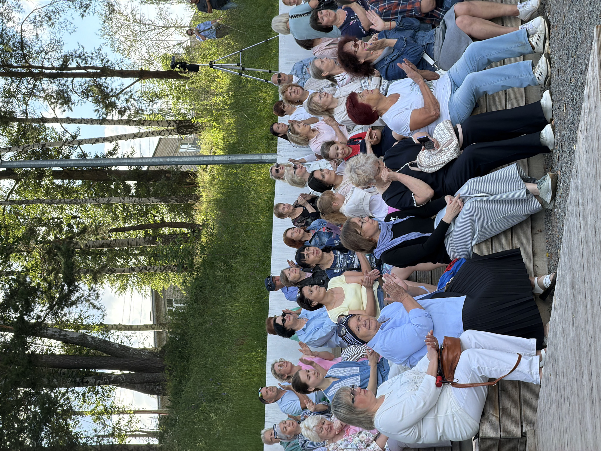 The image depicts an outdoor gathering of people, likely an audience, seated on tiered wooden benches. The setting appears to be a park or an open-air venue, with trees and a grassy slope visible in the background. A building with a brick facade and windows is situated on the slope, and in the distance, a person stands near a tripod-mounted camera. The audience members, primarily women of various ages, are dressed in casual attire. Some are clapping, suggesting they are enjoying a performance or presentation. The overall atmosphere is relaxed and social. The location is identified as Chaikovsky, Russia, possibly at an event or festival held in a public space within the city.