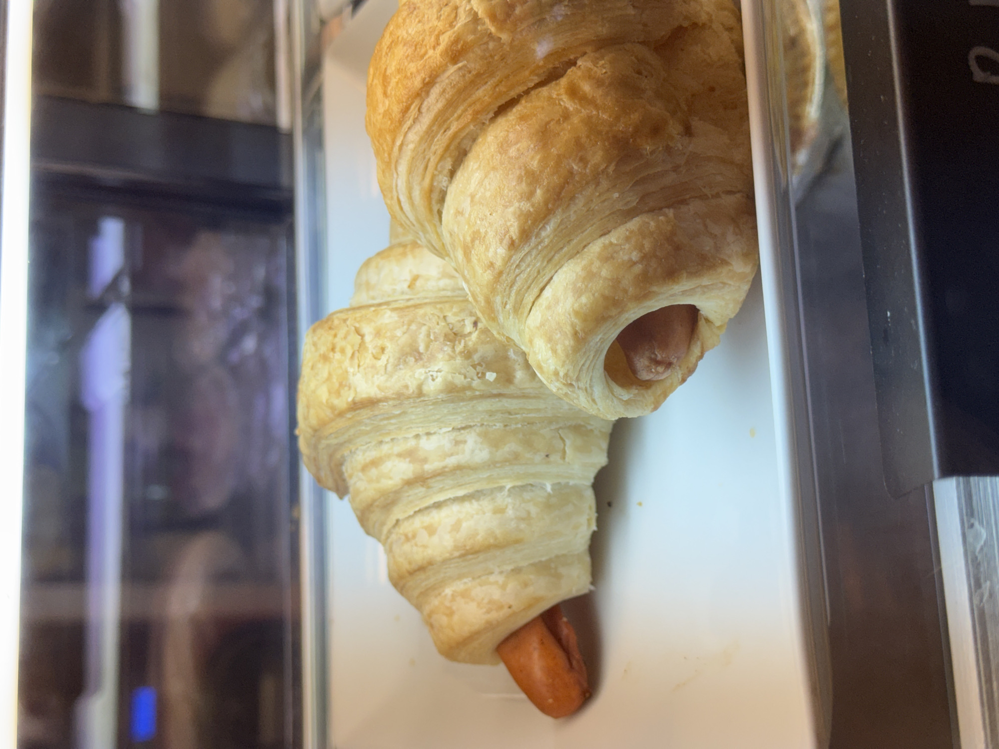 Two pigs in a blanket are displayed in a white rectangular dish within a glass-enclosed refrigerated display case. The croissants are golden brown and flaky, with a visible hot dog peeking out of one end of each. The background is blurred, showing reflections of lights and potentially other food items. The scene appears to be in a food establishment, possibly a bakery or cafe, in a commercial area.