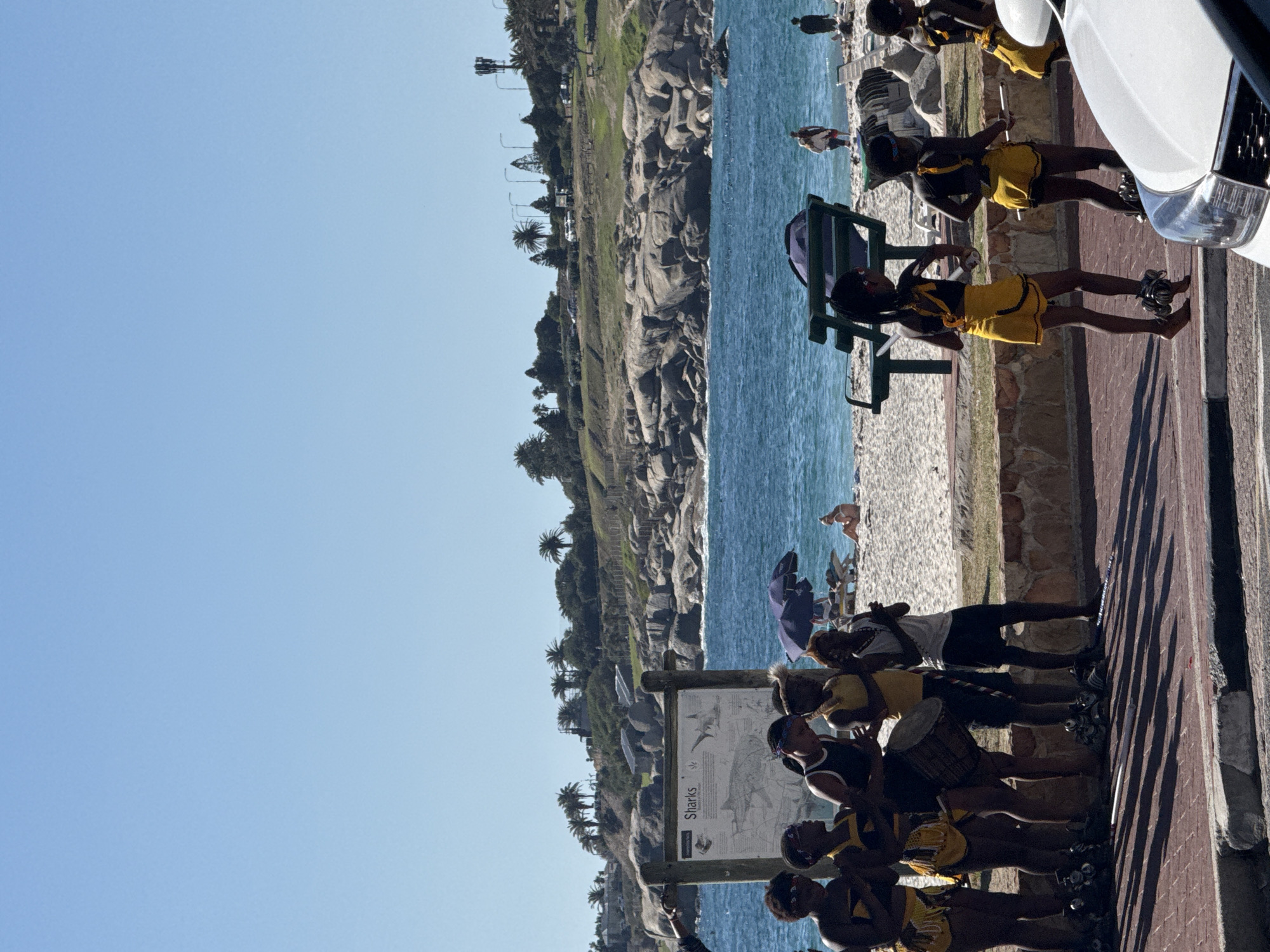 A landscape photograph of a group of African women standing next to the sea in Cape Town, South Africa, with the image rotated 90 degrees clockwise. The foreground features a brick walkway and the side of a white vehicle on the right side. The group of women is positioned along the walkway, some of them holding drums, while others are standing near a sign describing sharks. In the background, a beach stretches along the water's edge, dotted with beachgoers and umbrellas. Beyond the beach, a rocky coastline is visible, covered in vegetation. The sky above is clear and blue.