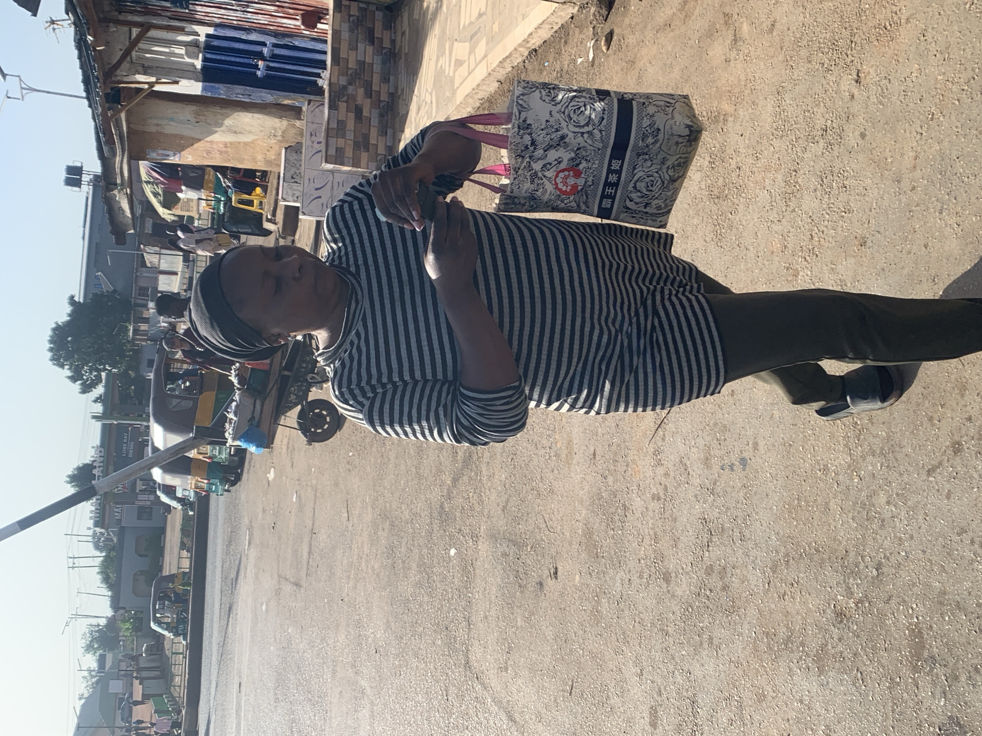 In the city of Gura Top, Nigeria, a woman wearing a black headscarf and a striped long-sleeved shirt walks on a dusty road. She is holding a patterned tote bag with pink straps in her right hand and appears to be interacting with her mobile phone in her left hand. The road is paved with asphalt and gravel. In the background, there are several buildings, including a row of shops with corrugated metal roofs. Various vehicles, including a tuk-tuk and a cart, are visible on the side of the road. The scene appears to be a busy commercial area on a sunny day, as indicated by the clear sky and the shadows cast by the objects.
