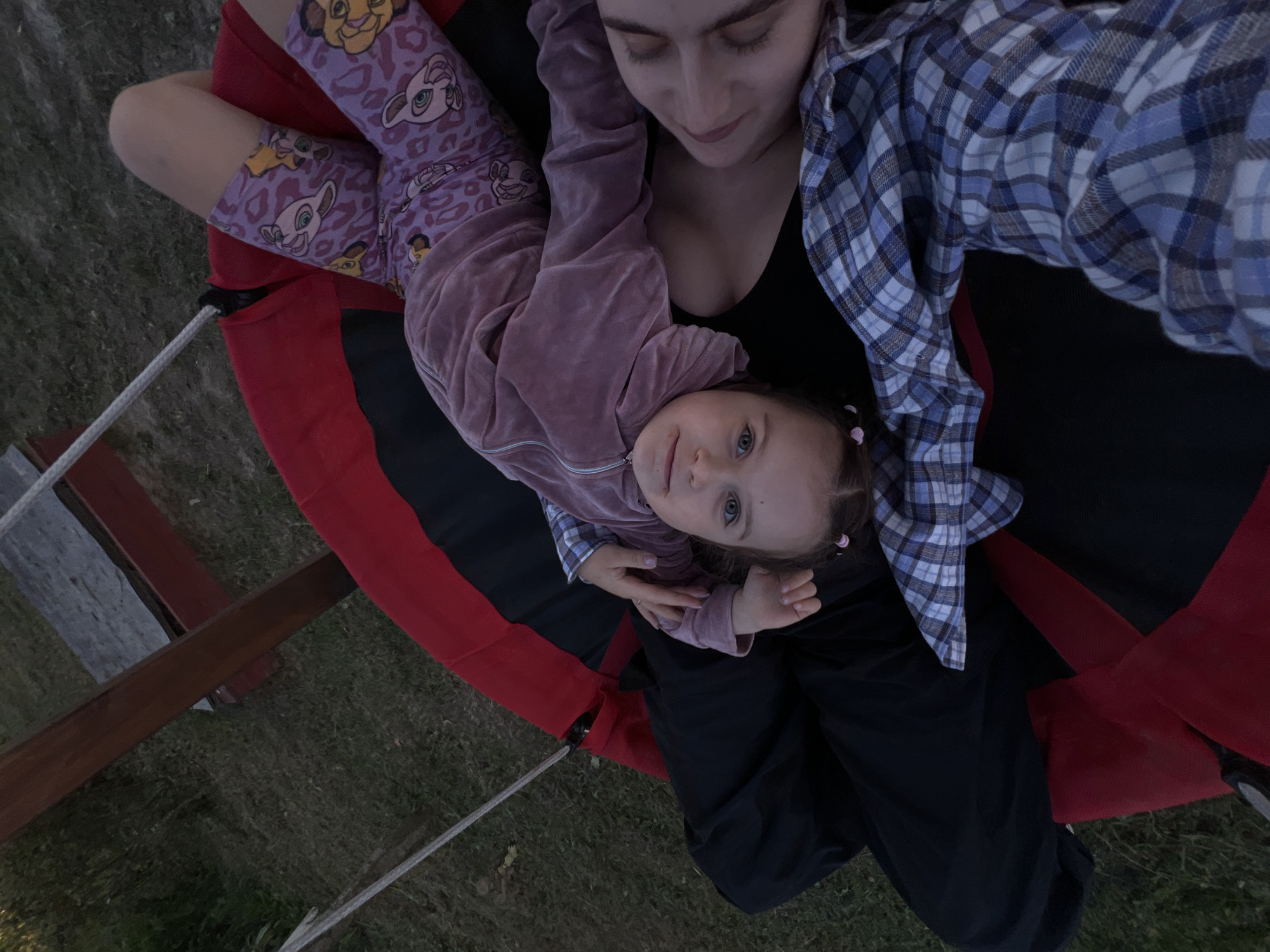 An overhead, high-angle, medium shot captures a woman and a young child lying on a large, round swing suspended from a wooden frame outdoors. The swing has a red exterior and a black interior. The woman is lying on her back, wearing a black tank top and a blue and white plaid shirt. Her head is tilted back and her eyes are closed. The child is lying on top of the woman, facing the camera with their head tilted to the left. The child is wearing a light pink velour hooded jacket and purple leggings with a pattern of lion cubs. The child has light brown hair with small pink hair clips. The background consists of green grass and a wooden structure. The lighting suggests it is late afternoon or early evening. The location is not identifiable as the city of Lublin, Poland, from the visual information provided in the image.
