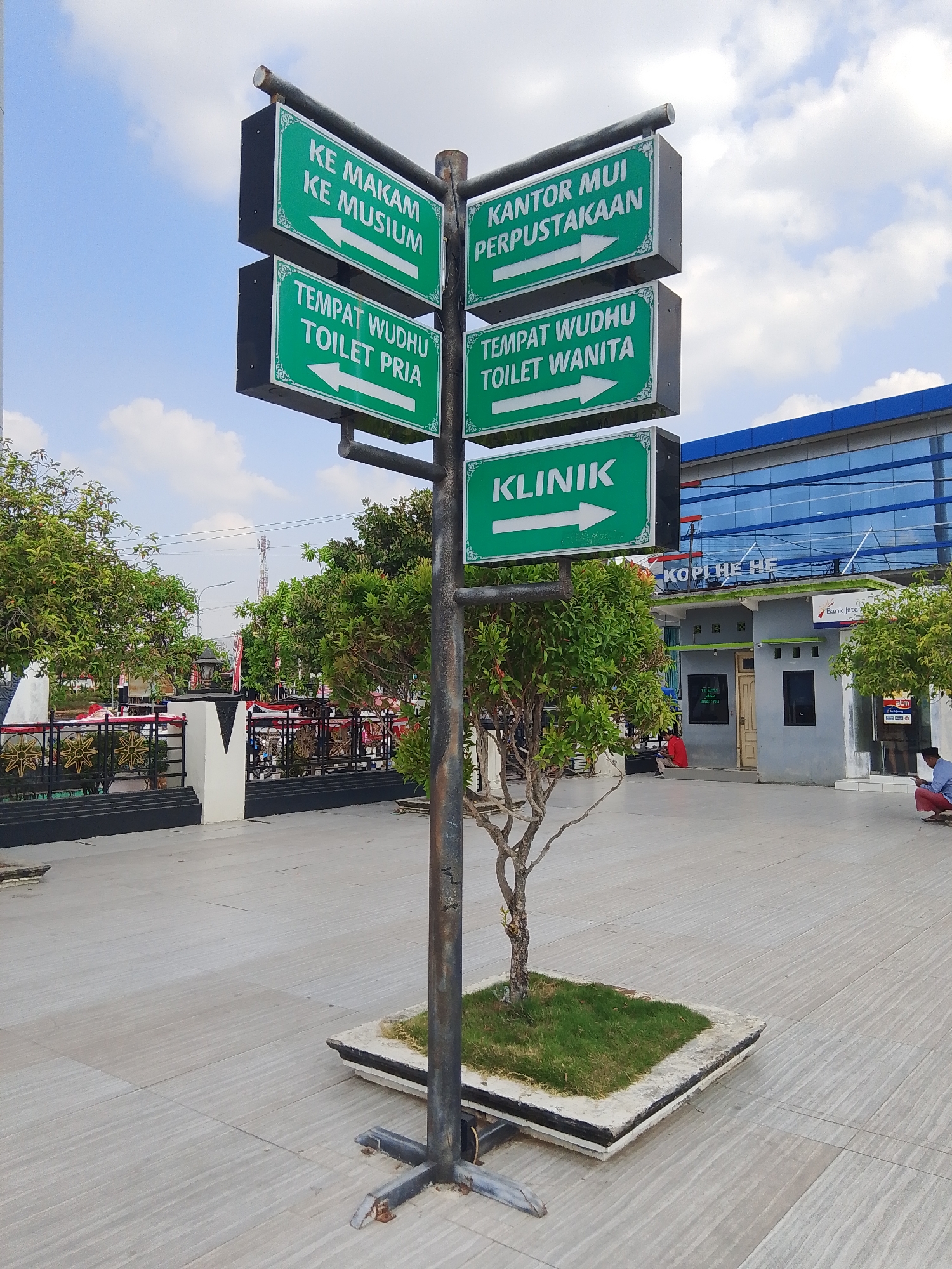 In Demak, Indonesia, a signpost with multiple green directional signs stands prominently on a paved outdoor area. The signs, written in white text, point towards "KE MAKAM KE MUSIUM" (To Cemetery, To Museum), "TEMPAT WUDHU TOILET PRIA" (Wudu Place, Men's Toilet), "KANTOR MUI PERPUSTAKAAN" (MUI Office, Library), "TEMPAT WUDHU TOILET WANITA" (Wudu Place, Women's Toilet), and "KLINIK" (Clinic). The signpost is anchored in a square planter with grass. In the background, a modern building with blue glass windows is visible, with a smaller building below it. A man in red clothing sits near the smaller building, and another man in a white shirt and red pants, wearing a black cap, is seated on the right side of the image, looking at a phone. Trees are present on the left and right sides of the scene. The sky is partly cloudy.