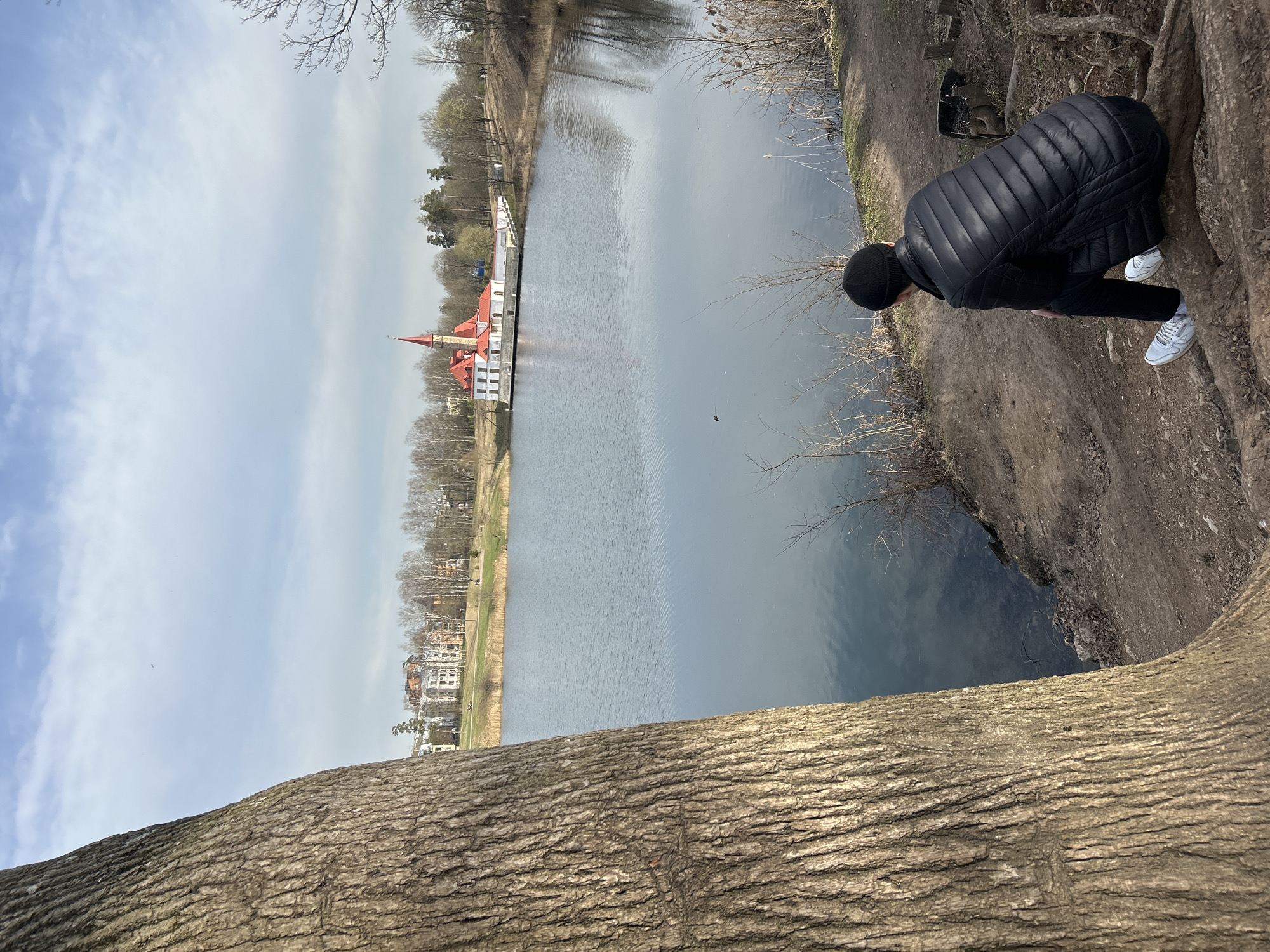 A ground-level shot of a man leaning over a tree trunk, looking towards a body of water. The man is wearing a black puffy jacket, black pants, a black beanie, and white sneakers. Across the water, there is a building with a red roof and white walls. Beyond the building, there are residential buildings and trees. The sky is partly cloudy. Location: Unknown, Unknown.