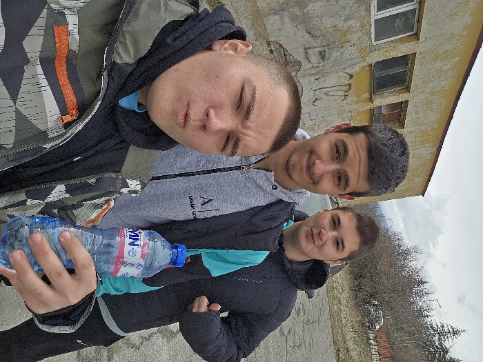 This image captures three young adult males in Samokov, Bulgaria, under an overcast sky.
The subject in the immediate foreground, positioned on the left, is male, with very short dark hair and light skin. He is wearing a multi-colored camouflage-patterned jacket over a dark hooded sweatshirt and a light blue t-shirt. His lips are pursed forward, and he holds a translucent blue plastic bottle with a red cap, branded 'DEVIN,' in his right hand. This individual is holding the camera, indicating a selfie.
To the right and slightly behind the foreground subject is a second male, with short dark hair and light skin. He is wearing a grey zip-up hoodie featuring a dark 'ARMANI EXCHANGE' logo on the left chest. His facial expression is a slight smile.
Further to the right and slightly behind the second subject is a third male, also with short dark hair and light skin. He is wearing a dark, possibly black, quilted jacket over a bright turquoise long-sleeved shirt. His right hand is raised in a 'thumbs up' gesture, and he exhibits a slight smile.
The background to the left features a two-story building with worn, faded yellow stucco and exposed brickwork, displaying graffiti, including letters resembling "BAR," on a lower wall section. Multiple rectangular windows with dark frames are visible on the building's facade. The ground surface in the middle ground appears to be asphalt or concrete. Beyond this, the distant background reveals a landscape with sparse, bare deciduous trees and shrubs. A dark-colored vehicle is partially visible among the vegetation on the right side, and distant hills or mountains are discernible beneath the cloudy horizon.