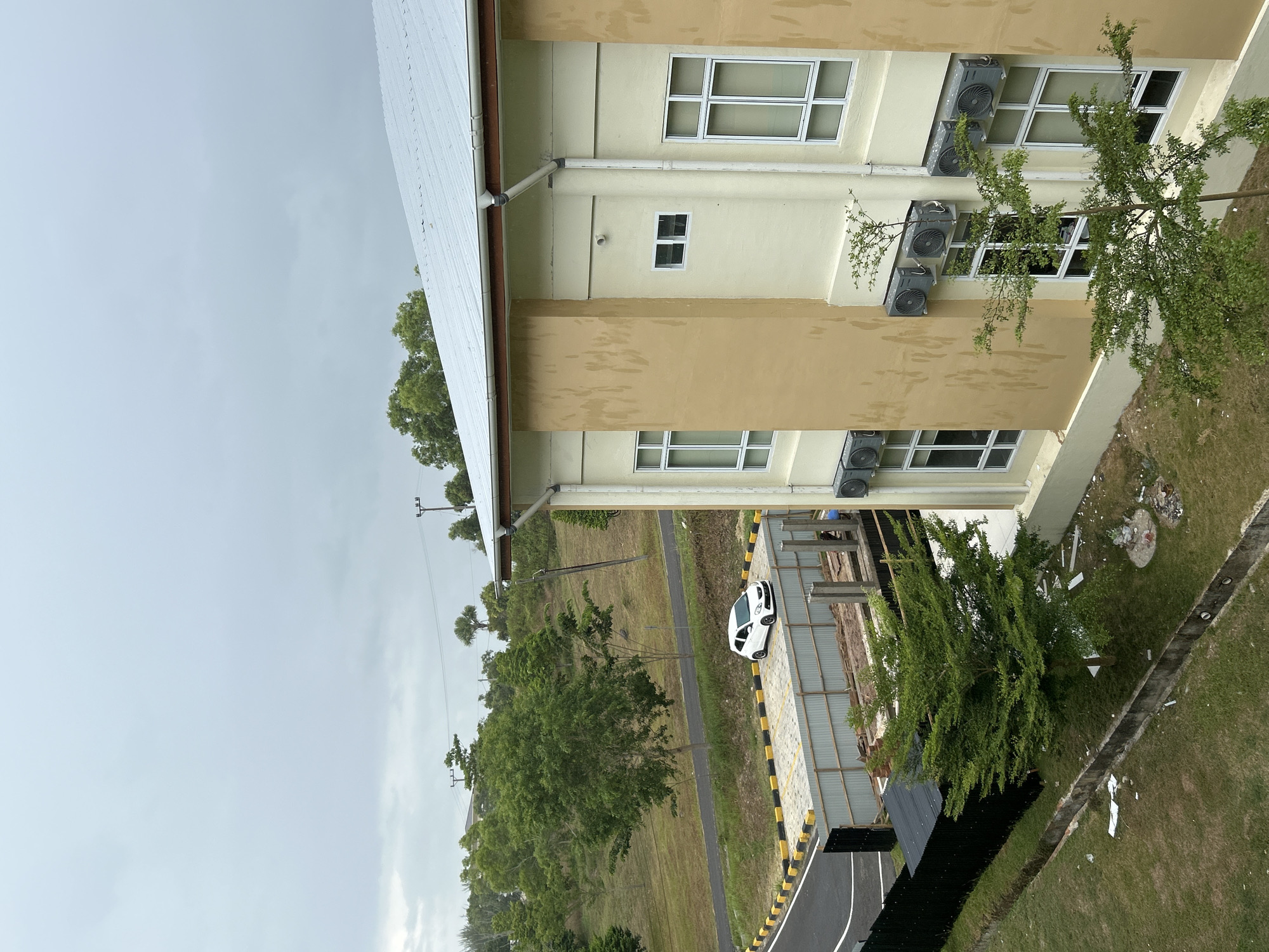 The image captures a daytime scene in Reuleut Barat, Indonesia, under an overcast sky. A multi-story building, likely an institutional or commercial structure, dominates the right side of the frame. The building features light yellow and brown painted walls, with multiple rectangular windows fitted with white frames. Several grey external air conditioning units are mounted below the windows on the building's exterior. A white gutter system runs along the roofline.
In the mid-ground, a paved road with black and yellow striped curbing and white lane markings is visible. A white car with a black roof is parked on a paved area adjacent to the road, near the building. Beyond the road, the landscape consists of a vast expanse of green, gently rolling terrain, dotted with numerous trees and some utility poles stretching towards the horizon.
Closer to the building, there's a patch of green grass with some scattered debris, including what appears to be discarded plastic and construction materials. A dark, possibly metal, fence or wall is present in the lower foreground. No people or specific activities are visible in the scene. The uniform light grey sky suggests a cloudy or overcast day. No visible text is present in the image.