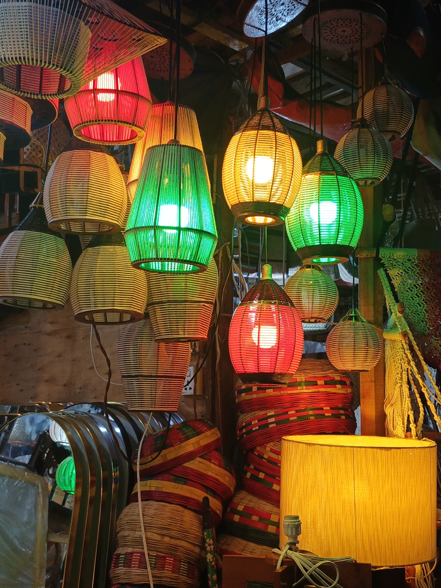 This image captures a vibrant and densely packed display of handcrafted lamps and woven decorative items, likely in a market stall or shop in Dhaka, Bangladesh. The primary subjects are numerous ceiling-mounted pendant lamps and one large table lamp, showcasing diverse shapes and vivid colors.
The hanging lamps vary in form, including spherical, cylindrical, and conical designs, all appearing to be intricately woven from natural materials like bamboo, rattan, or straw. Many are illuminated from within, casting bright red, green, and yellow (or amber) light that creates a warm and inviting glow. The textures of the woven materials are clearly visible, with some lamps featuring horizontal bands of weaving and others vertical strips, highlighting the artisanal craftsmanship.
In the bottom right, a prominent drum-shaped table lamp with a yellow fabric shade adds to the illumination. Below the hanging lamps and around the base of the display, stacks of colorful woven baskets or containers are visible, adorned with intricate patterns of red, green, and yellow, complementing the color scheme of the lamps. Various wires and electrical components are discernible, connecting the light fixtures. Metallic or reflective surfaces are partially visible on the bottom left, suggesting other wares or display elements.
There are no people visible, and no specific activity is taking place beyond the static display of merchandise. The reliance on artificial light from the lamps themselves suggests an indoor setting, possibly during evening hours or within a space with limited natural light. The abundance of handcrafted, colorful items evokes a lively market atmosphere common in South Asia. No legible text is visible in the frame.