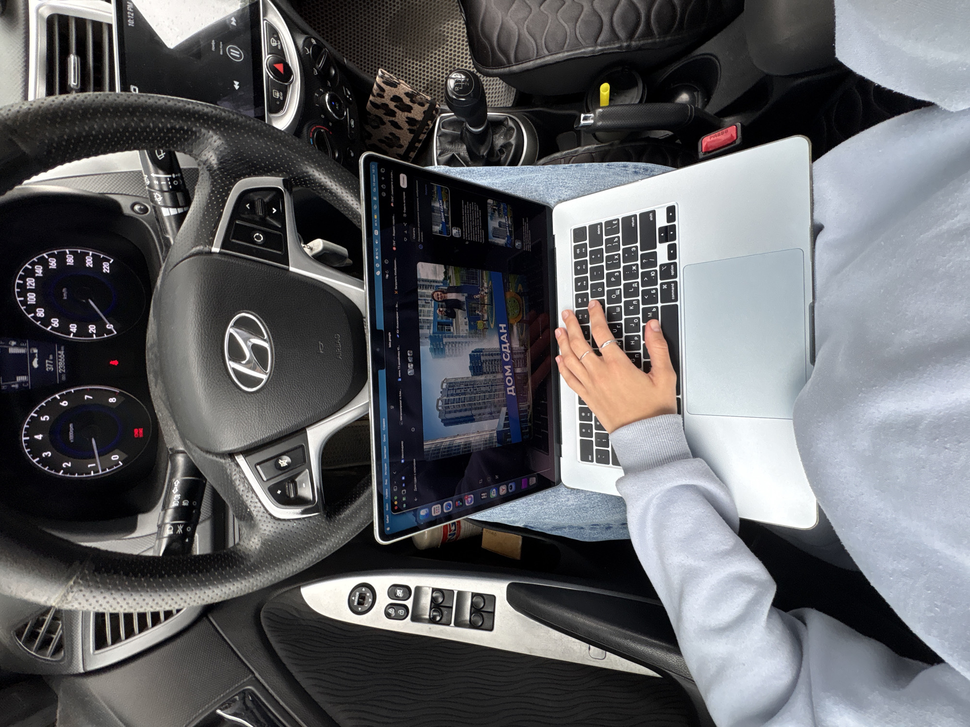 In a parked car, a person wearing a light blue sweatshirt and denim jeans is seated in the driver's seat, working on a silver laptop. The laptop is open and placed on their lap, with their right hand typing on the keyboard. The car's interior is visible, including the steering wheel with a Hyundai logo, the dashboard with gauges and a car stereo screen, and the gear shift. A leopard-print wallet is placed on the passenger seat. The image appears to be taken from a high angle, looking down at the person and the car's interior. The context of this image is not specific to the city of Perm, Russia, as no identifying landmarks or signs are present.