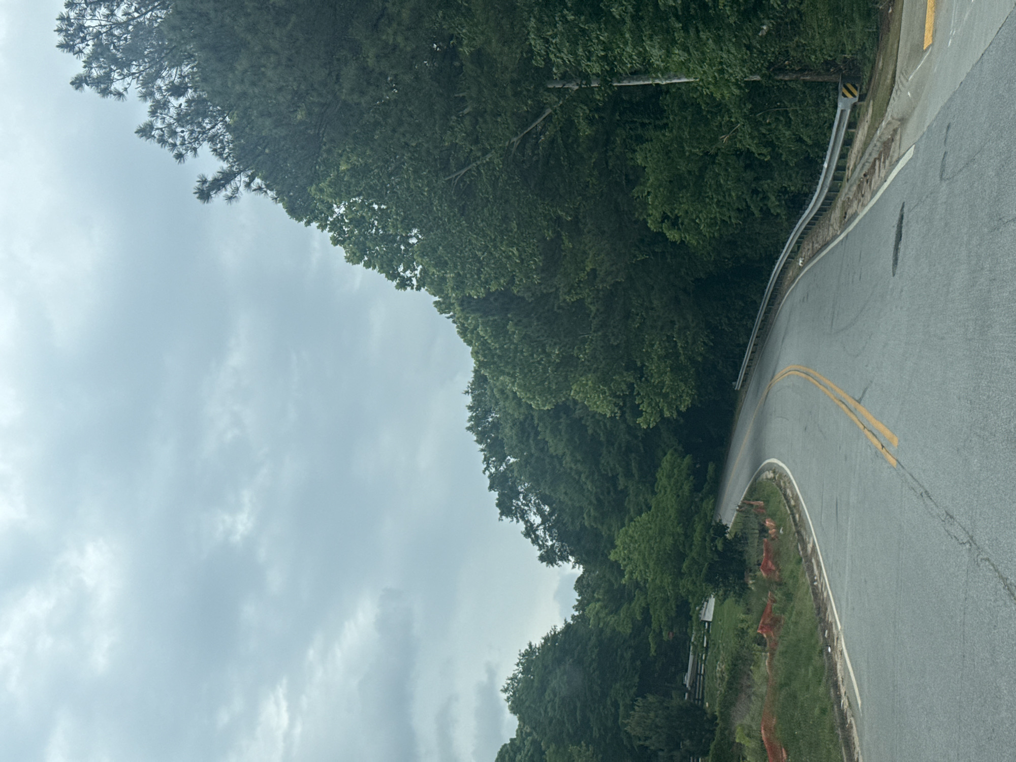 The image depicts a curving asphalt road in Mountain Park, United States of America. The road has double yellow lines indicating the center. A metal guardrail runs along the side of the road. Trees densely border the road. The sky is overcast with clouds.