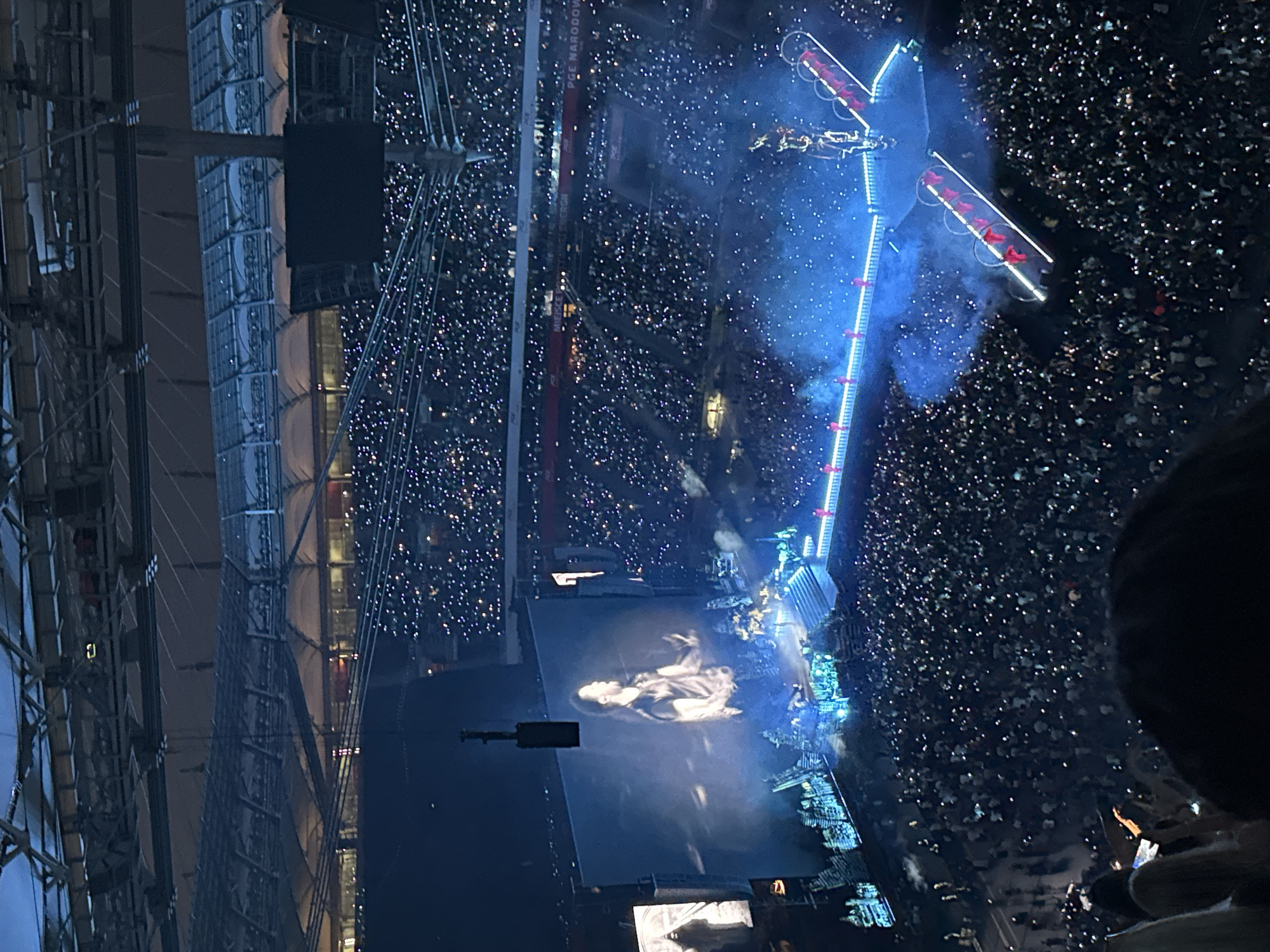 The image depicts an aerial view of a large stadium concert, likely held at PGE Narodowy in Warsaw, Poland. The stadium is filled with a vast audience, with many individuals holding up illuminated mobile phones, creating a sea of white lights. A prominent, large screen displays a live projection of a male performer on stage. The stage itself is elaborate, featuring a walkway with bright blue lighting and several individuals in red attire visible on elevated platforms. Smoke or fog effects are present on the stage, adding to the atmospheric production. The stadium's roof structure is visible, highlighting the indoor/outdoor nature of the venue. The overall scene captures a dynamic and energetic live musical event.